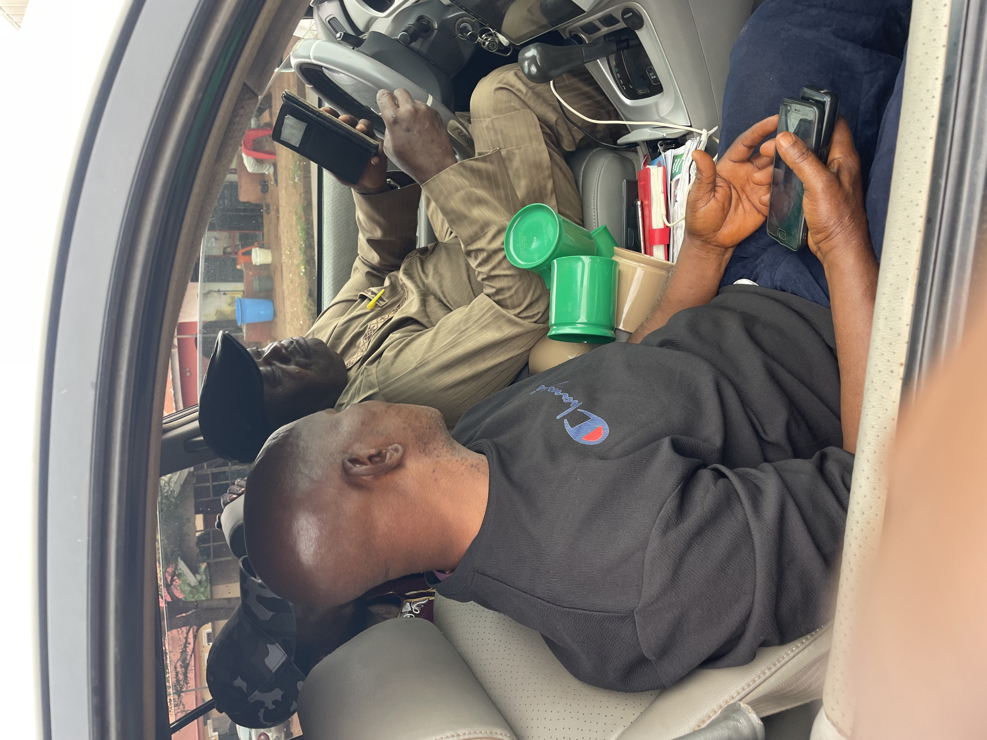 Two men are sitting inside a vehicle, likely a car, in Okpo, Nigeria. The man in the driver's seat is wearing a tan uniform and a black baseball cap. He is looking at a black mobile phone in his hands. The passenger is wearing a black Champion sweatshirt and a black baseball cap with a grey camouflage pattern. He is looking at two phones in his hands. Several green plastic cups are positioned between the two men, along with several notebooks and a phone charger. The interior of the car is light grey leather. The car window shows the reflection of the outside with some red-bricked and off-white-colored buildings.