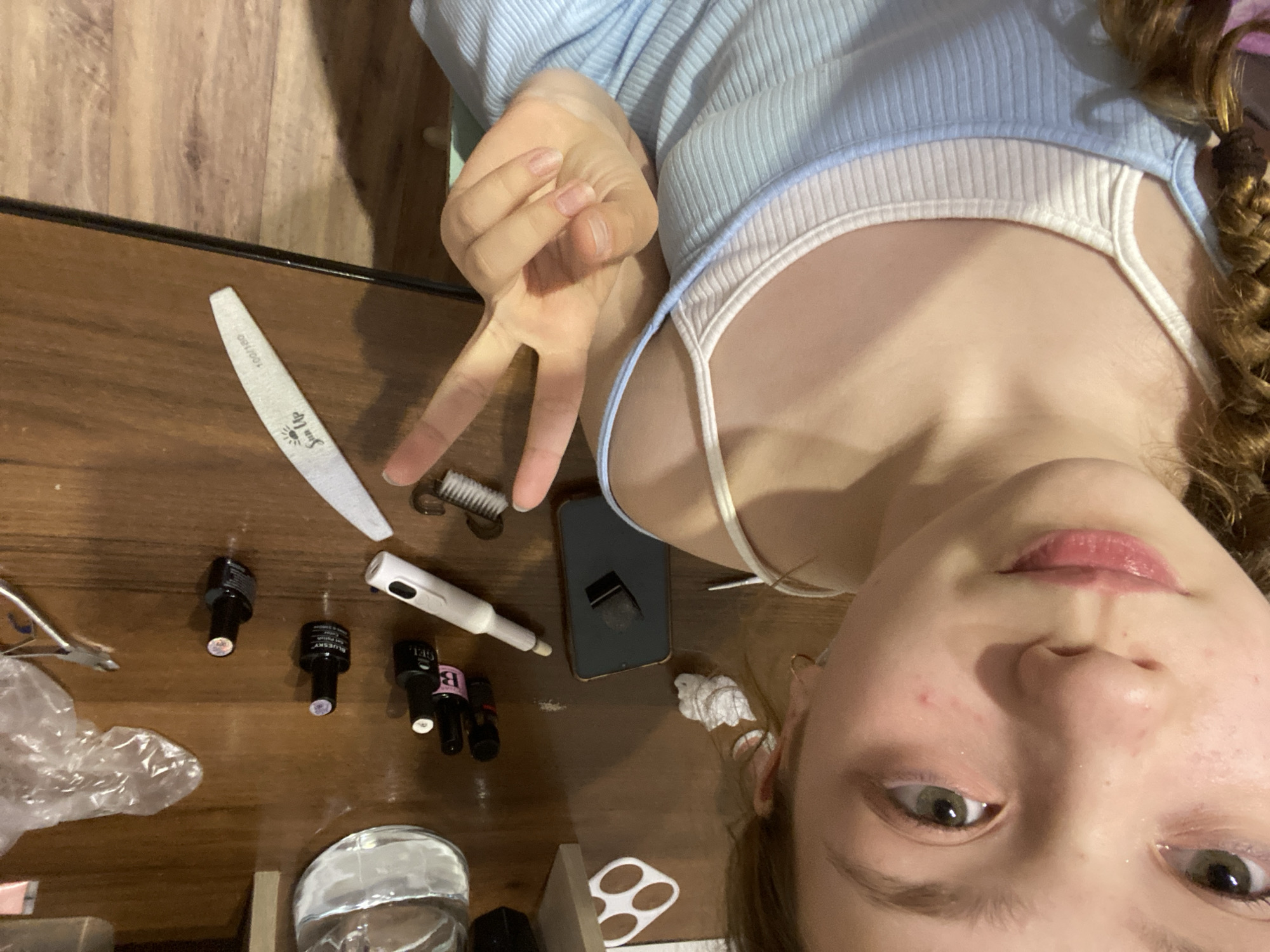 A selfie photograph captures a young girl with light skin and braided brown hair, wearing a light blue top over a white tank top. She is making a peace sign with her fingers. The background is a wooden surface upon which are several manicure-related items: a nail file, a brush, a phone, a nail drill pen, nail polish bottles, and a small container of water. The location is unknown.