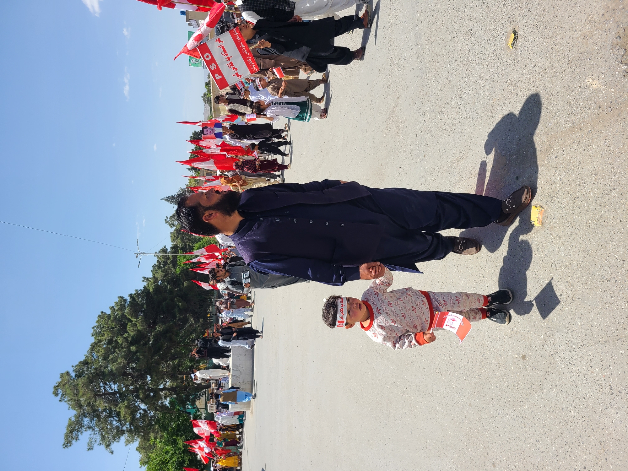 Here is a description of the image:
The image shows a street scene in Quetta City Tehsil, Pakistan, where a crowd has gathered for what appears to be a political demonstration or rally. Many people are visible in the background, holding red and white flags. A man in a dark blue traditional outfit holds hands with a child wearing an Adidas tracksuit and a headband that bears political text. The child is holding a small red and white flag. The scene is outdoors on a bright, sunny day, as evidenced by the clear sky and the shadows cast by the individuals. A large tree stands to the side of the street. The ground is paved.