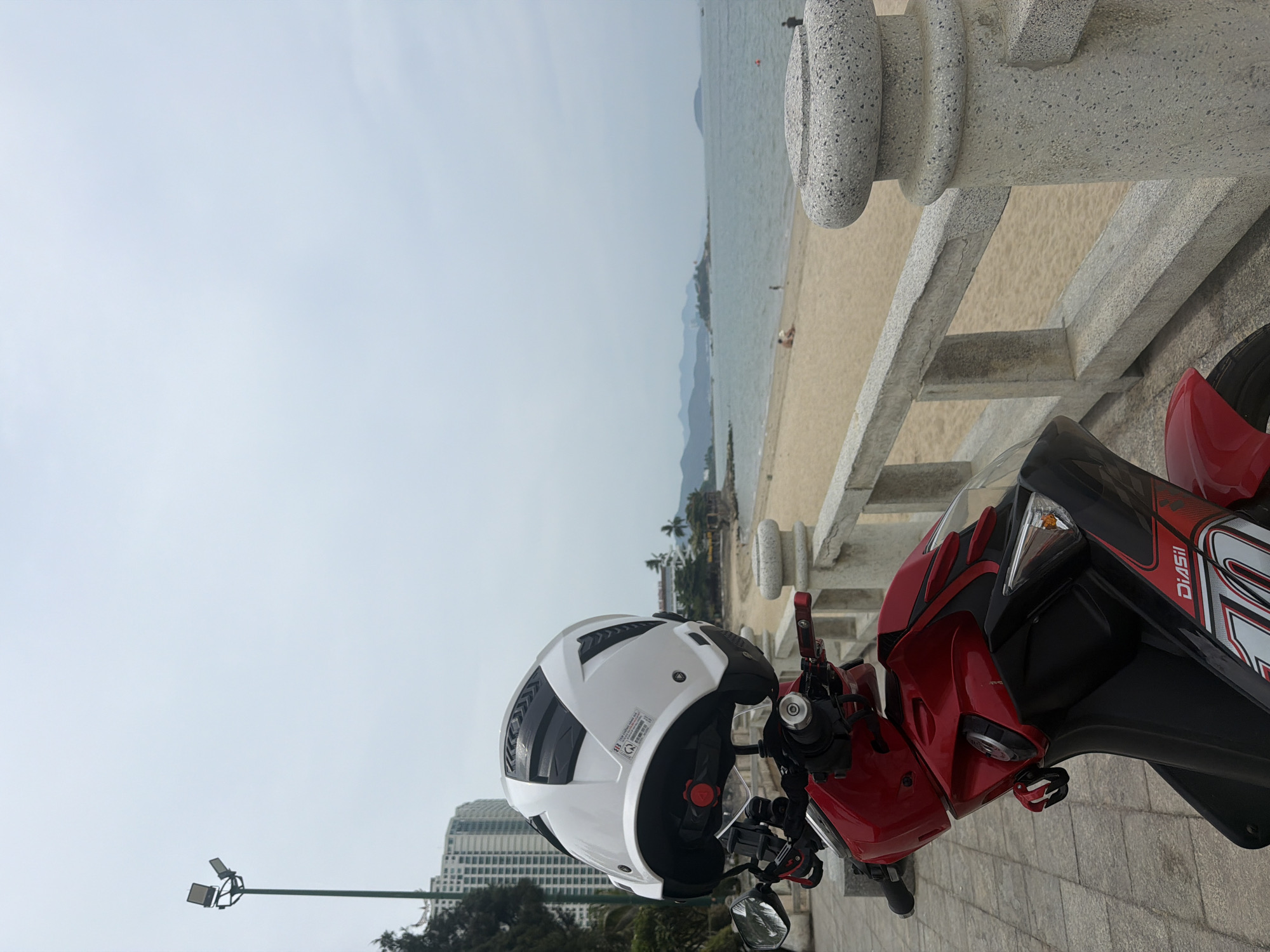 The image depicts a white motorcycle helmet resting on the handlebars of a red motorbike. The motorbike is parked next to a concrete railing. In the background is a beach with mountains in the distance, and a building on the left. The scene is in Nha Trang Ward, Vietnam.