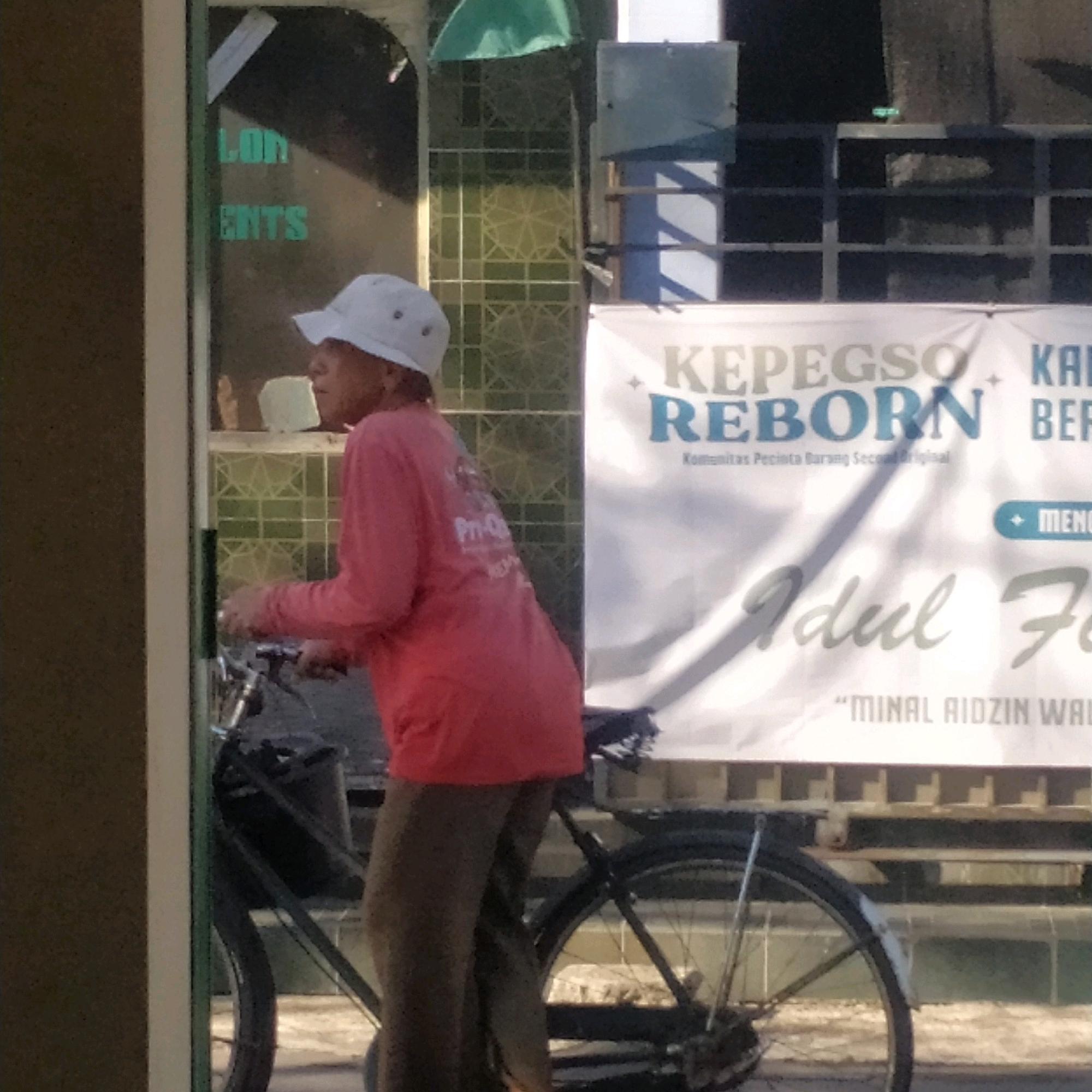 In Batan, Indonesia, a person wearing a white hat and a pink long-sleeved shirt rides a bicycle. The individual is positioned in the foreground, with their profile facing left. Behind them, a white banner displays text, including "KEPEGSO REBORN" and "MINAL AIDZIN WAL." A green tarp hangs in the upper left quadrant of the image, partially obscuring the background. The scene appears to be outdoors, with a building visible on the left side of the frame.