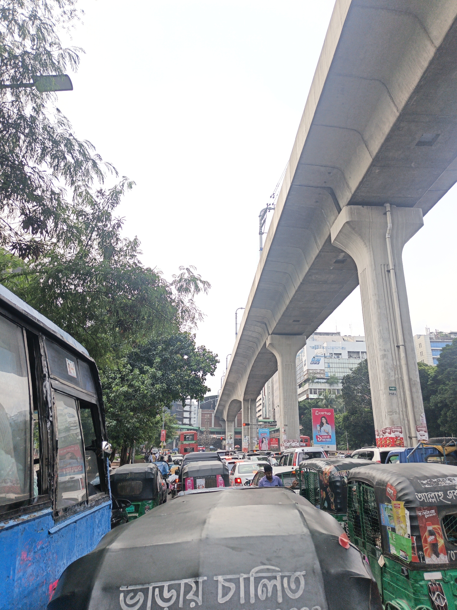 This image captures a street scene in Dhaka, Bangladesh, featuring heavy traffic congestion beneath a prominent elevated road or flyover. A large blue bus is on the left, its windows reflecting the surrounding environment. Several auto-rickshaws and cars are visible, bumper-to-bumper, indicating a significant traffic jam. Trees line the side of the road, and buildings can be seen in the background, some with advertisements. The sky is overcast. The elevated structure dominates the upper portion of the image, supported by large concrete pillars. The overall impression is one of urban hustle and bustle, characteristic of Dhaka's busy thoroughfares.