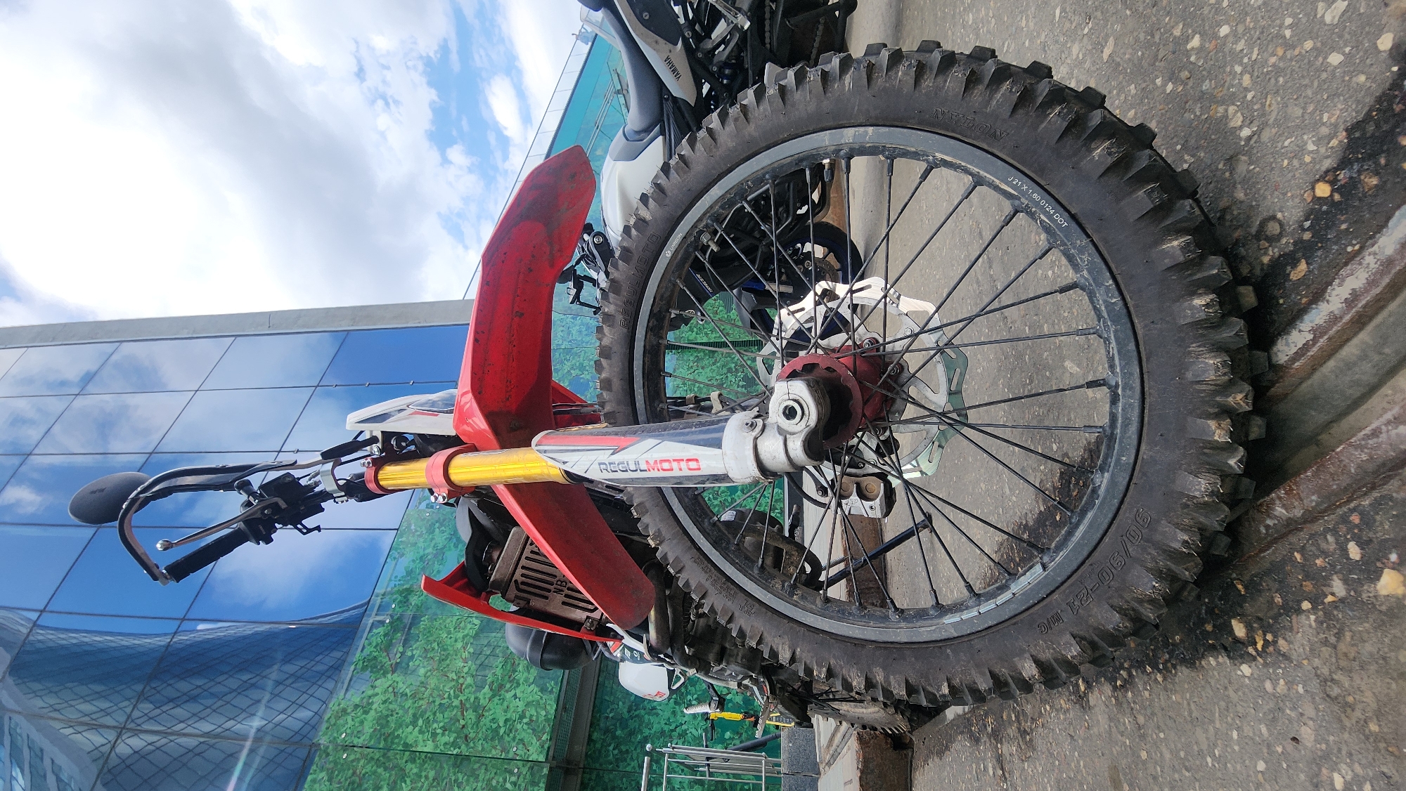 The image shows a close-up of the front wheel of a red and white Regulmoto motorcycle. The tire is a knobby off-road type with the size "J 21 X 1.60 0124 DOT" and "90/90-21 M/C" printed on its sidewall. The front fender is red. The motorcycle is parked on a paved surface. In the background, there's a modern building with a glass facade reflecting the sky and clouds. Another motorcycle, a Yamaha, is partially visible behind the Regulmoto. The location is Moscow, Russia, judging by the Cyrillic characters on the Yamaha motorcycle.