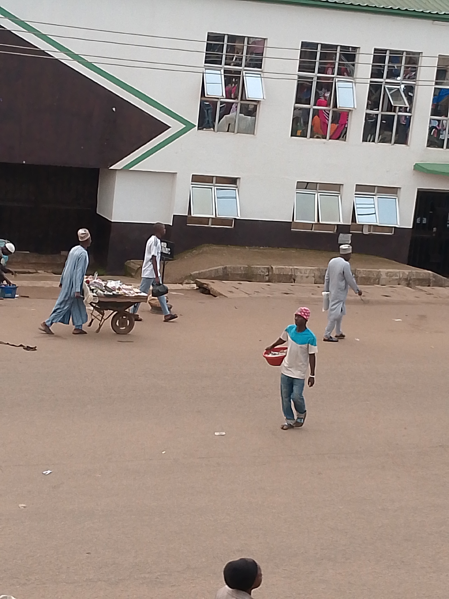 The image captures a street scene in Bukuru, Nigeria, with a focus on pedestrian activity and the architecture of a building in the background. A cream-colored building with multiple windows dominates the upper portion of the frame. Some windows are open, revealing glimpses of interior spaces with what appears to be clothing or fabrics. The building features a dark brown section with a distinctive green and white diagonal stripe design.
In the foreground, several individuals are present. One person, dressed in a light blue traditional garment and a white cap, is pushing a cart laden with goods, possibly merchandise for sale. Another person, in a white t-shirt and jeans, is walking behind the cart, carrying a dark bag. Further to the right, a person wearing a light grey traditional outfit and a cap walks away from the viewer. In the lower right quadrant, a person in a white and blue t-shirt and jeans, with a pink and white head covering, carries a red bowl filled with items. The ground is paved and appears to be a street or marketplace, with scattered litter. The overall atmosphere suggests a typical day in a bustling Nigerian city.