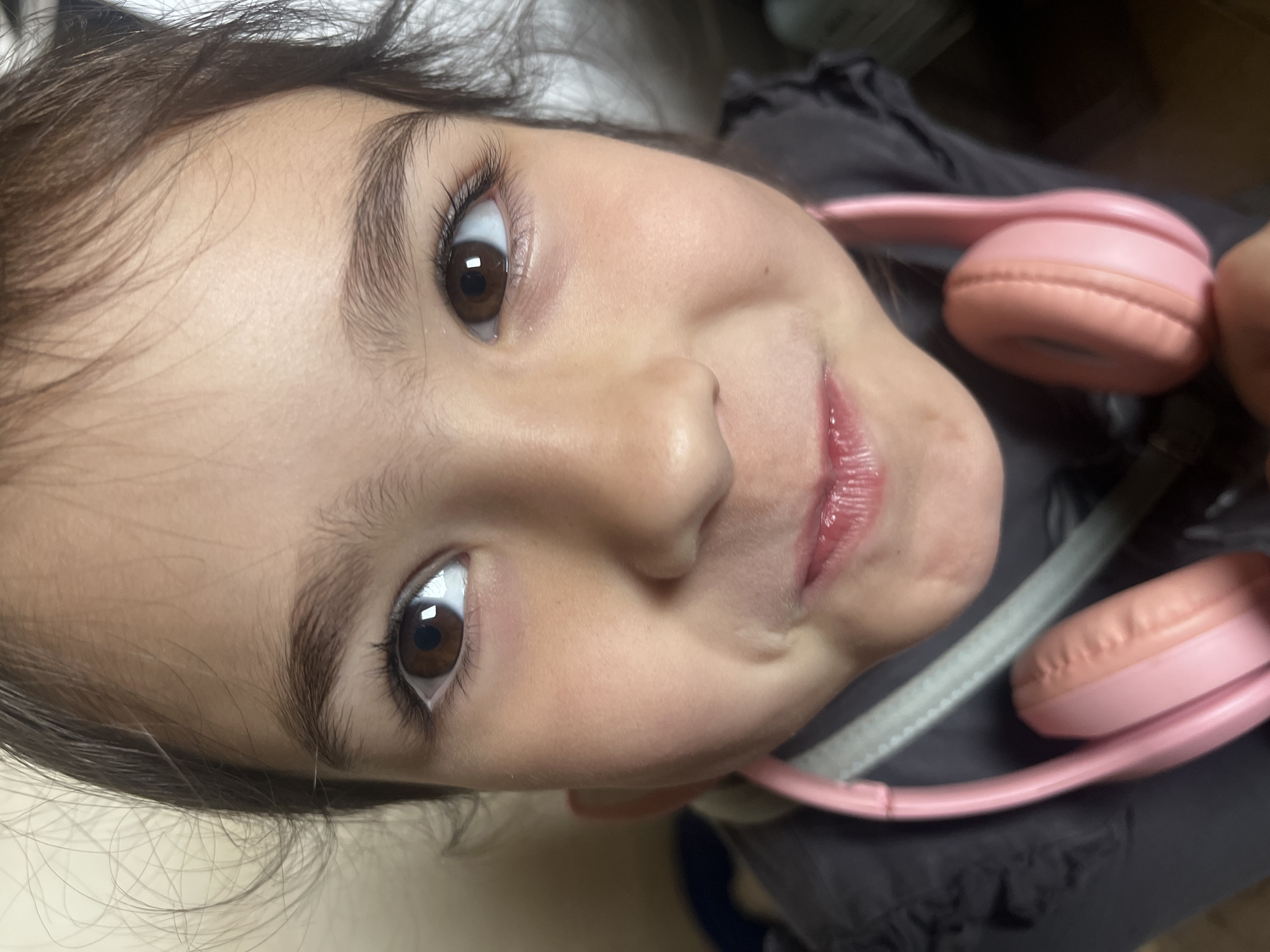 A close-up, angled shot depicts a young child's face with pink headphones draped around their neck. The child has brown eyes, dark hair, and fair skin. The location is not discernible from the image and no information is provided to confirm the location to be Onokivtsi, Ukraine.