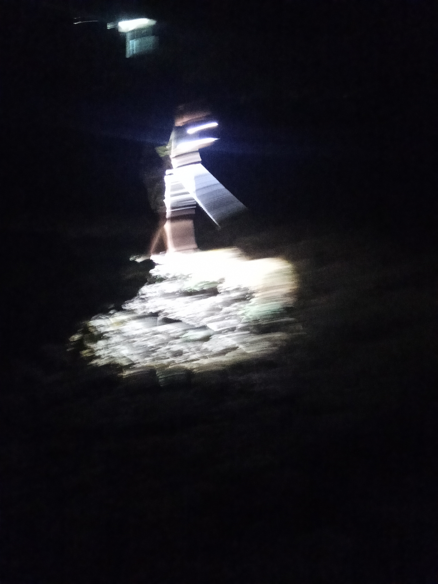 The image is a blurry, low-light photograph taken at night in what appears to be an outdoor setting, possibly a yard or pathway. A bright light source, possibly a flashlight or camera flash, illuminates a portion of the ground which is covered in rough, possibly rocky or uneven terrain. The light creates a strong glare and casts long shadows, indicating a dark environment with limited visibility. In the upper portion of the image, there is a suggestion of a structure, perhaps a building or a fence, with some light emanating from it as well. The overall impression is one of motion blur, as if the camera or the subjects were moving during the exposure. There is no discernible evidence within the image to suggest it was taken in Makale, Indonesia.