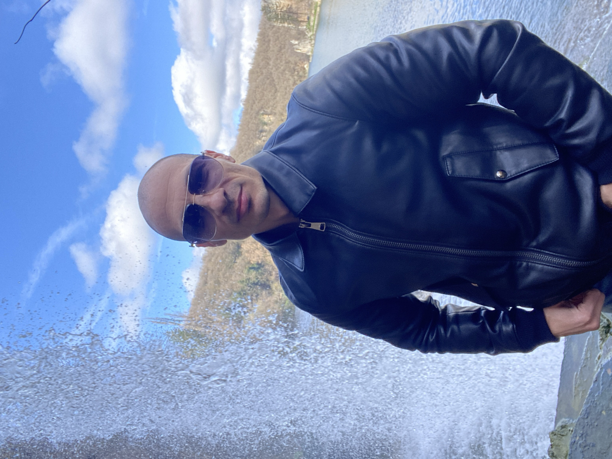 An adult male, bald and wearing sunglasses with a light-colored frame and dark lenses, stands centrally in the foreground. He is dressed in a dark, possibly black, leather jacket with a zippered front and a collar. A small flap pocket with a silver snap is visible on the jacket's right side. His hands are in his pockets, and he faces the camera with a neutral expression. To his immediate left, a large volume of water cascades downwards, creating significant spray. Behind and to his right, a body of water, appearing as a lake, extends into the midground. The background features rocky, barren hillsides with sparse, dry vegetation under a bright blue sky with scattered white cumulus clouds. The ground where the man stands consists of dark, irregular rock formations, possibly wet from the adjacent waterfall. The scene is consistent with the "Skoka" waterfall area within Kaylaka Park in Pleven, Bulgaria.