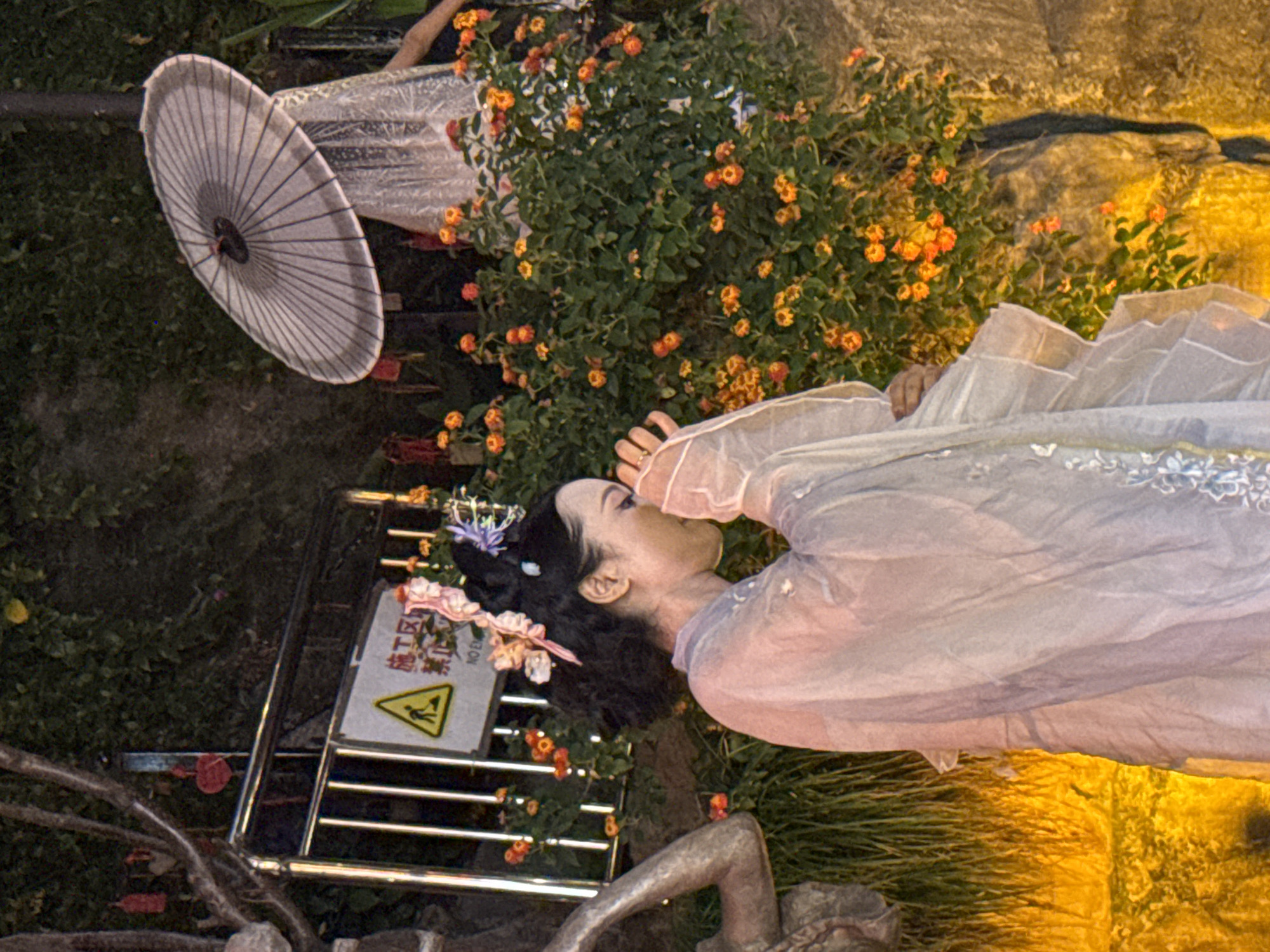 A woman in traditional Chinese attire, a light-colored layered robe and elaborate hair ornaments, stands with her head bowed and hands near her face, appearing to be in prayer or contemplation. She is positioned in front of a bush of small orange and yellow flowers. Behind her, a person partially obscured by a white umbrella and a lace veil is visible. In the background, a yellow hazard sign with Chinese characters indicating "construction area" and "no entry" is attached to a metal fence. The scene is lit with warm, artificial lighting, casting shadows and highlighting the textures of the foliage and the woman's costume. The environment appears to be outdoors, possibly a garden or a scenic area within Yuzhong District, China, given the presence of traditional elements and the context of a possible performance or cultural event.