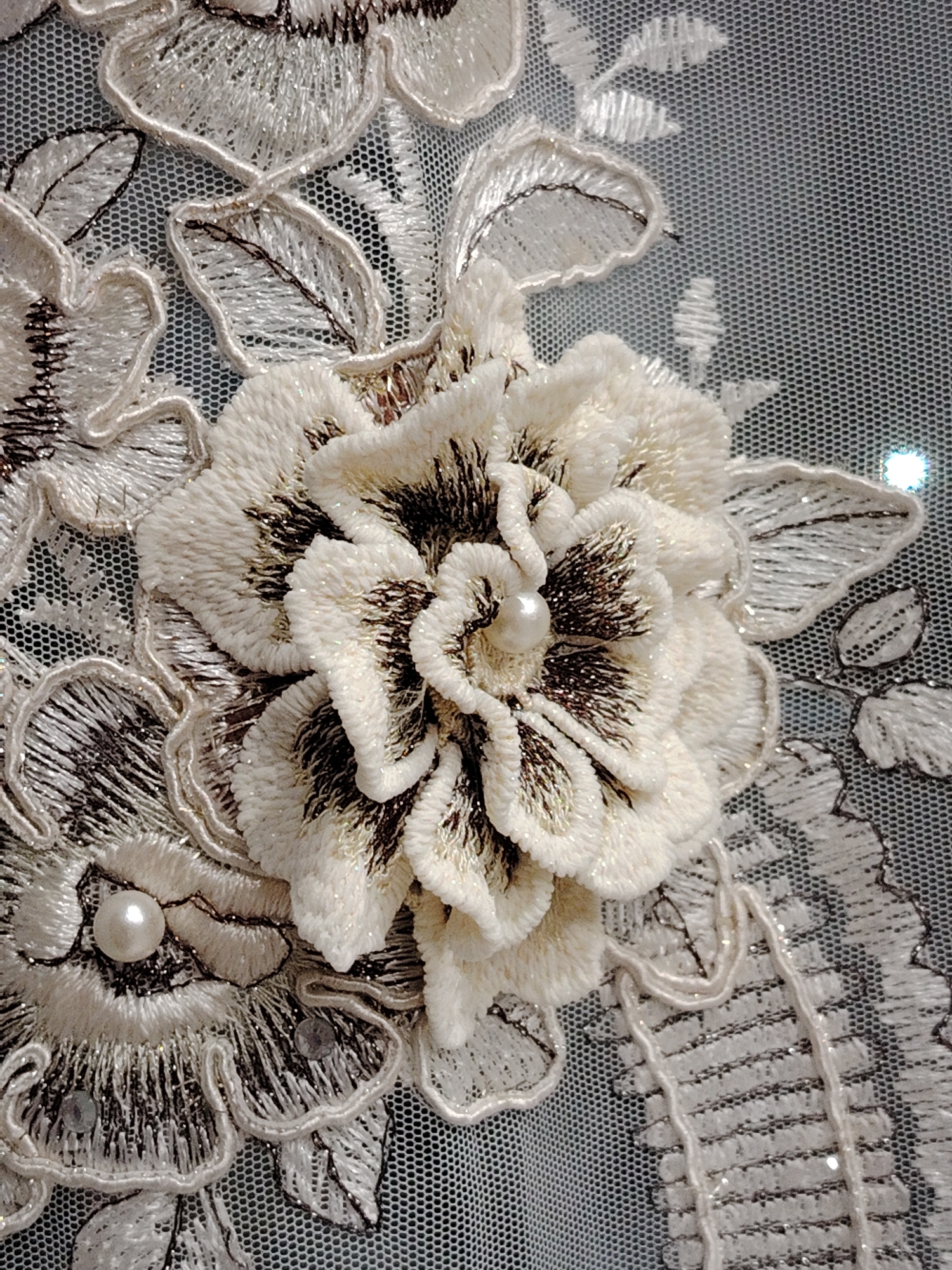 A close-up shot showcases an intricate piece of embroidered fabric, possibly from a wedding gown or similar formal attire. The fabric is a fine net, and the embroidery features several floral motifs in shades of white and cream. The central flower is particularly detailed, with layered petals and a pearl-like bead at its core. Darker stitching accents create shading and depth within the flower. Other floral designs and leaves, outlined with decorative stitching, surround the central flower. Small, shiny sequins are sparingly integrated into the design, adding a subtle sparkle. The background is slightly blurred, highlighting the texture of the net fabric.
This image is not associated with a specific city, especially not with Noviy, Russia. It's simply a macro photograph of an embroidered fabric sample.