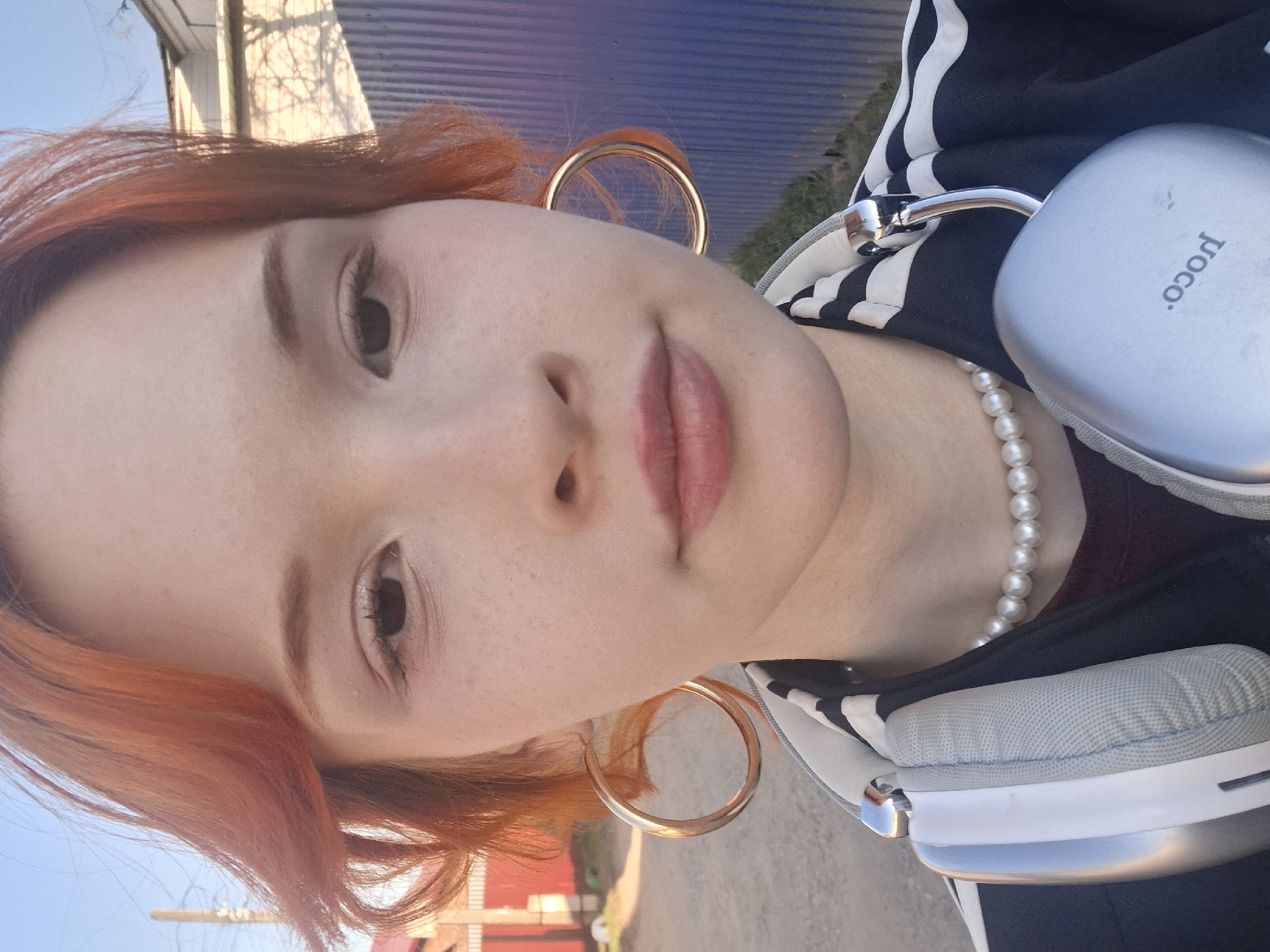 A close-up, angled selfie shows a light-skinned person with red-orange hair, wearing large gold hoop earrings, a pearl necklace, and a black and white striped jacket. Part of a grey "poco" brand headphone case is visible on the right side of the image, near the person's shoulder. The background includes glimpses of a building, possibly a fence, and the sky, suggesting an outdoor location. It is difficult to determine if the picture was taken in the city of Chaikovsky, Russia.