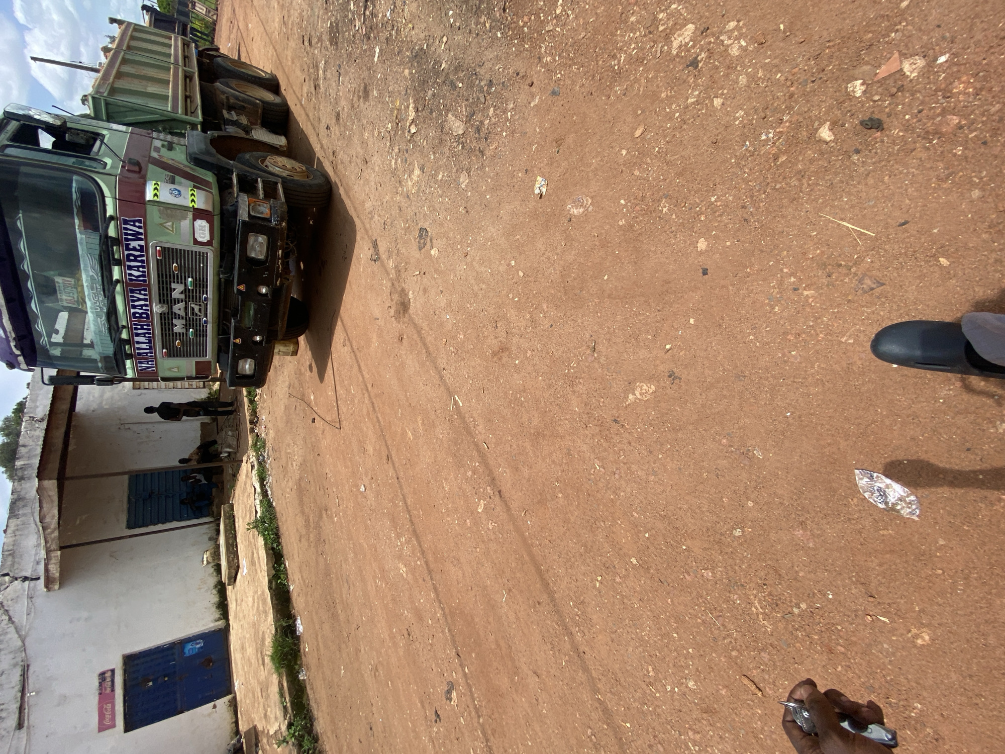 The image depicts a street scene in Gura Top, Nigeria. In the foreground, a person's hand holds keys, and their feet, clad in dark footwear, are visible. The ground is a dirt road with scattered debris. A large, green and maroon dump truck, identified with "MAN" on the grille and "NA ALLAH BAYA KAREWA" on its side, is parked on the right side of the road, loaded with sand. Behind the truck, a building with a white facade and corrugated metal roofing is visible. Two blue doorways are present, one with a Coca-Cola advertisement. Several individuals are gathered near the building, some sitting and one standing. The sky is partly cloudy.
