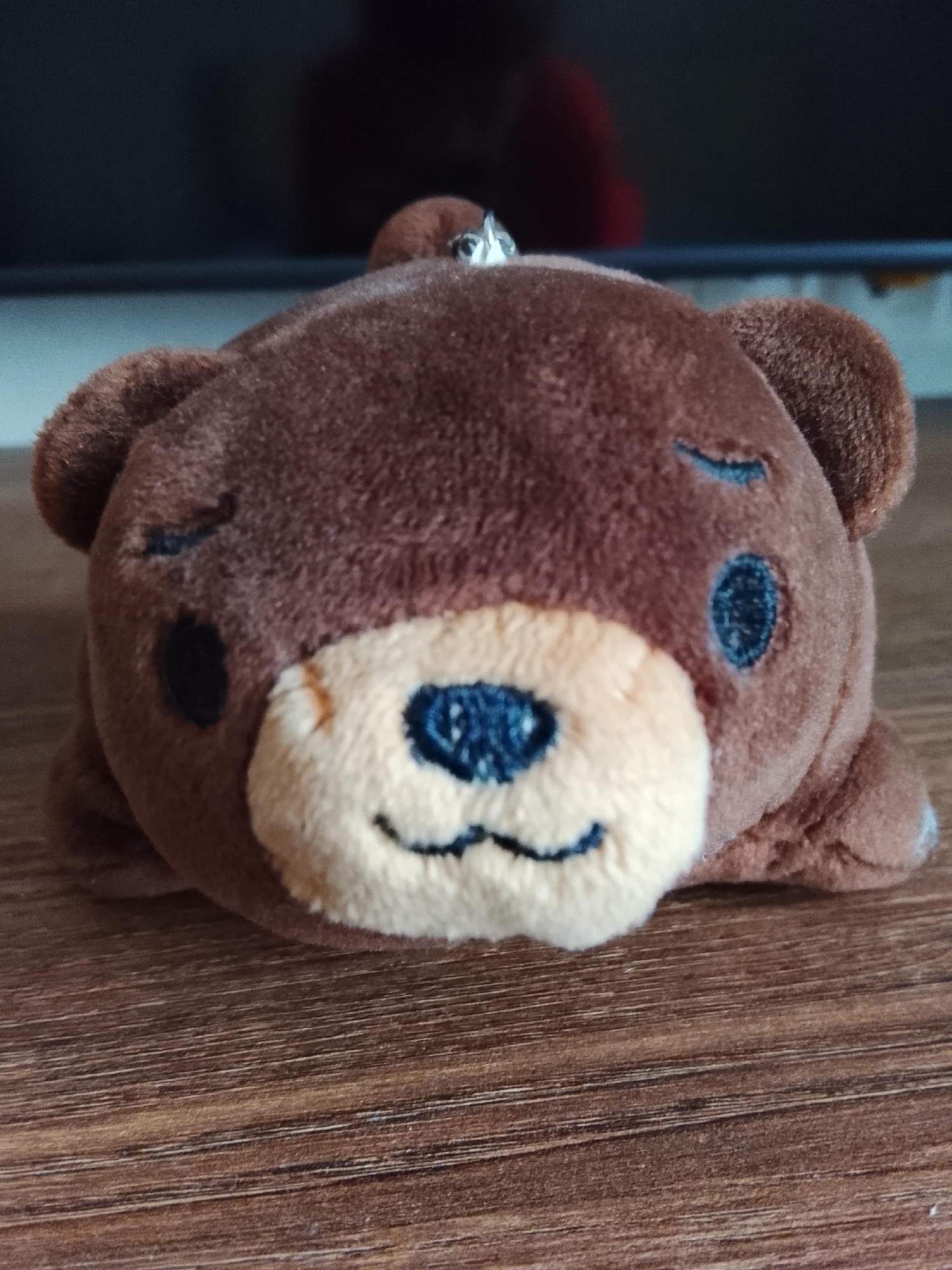 A close-up, eye-level shot shows a small, brown plush teddy bear lying face down on a wooden surface. The bear has closed eyes, a light tan muzzle with a stitched smile and nose, and black circular ears. A silver keychain loop is visible at the top of its head. The background is dark and out of focus, suggesting it is a flat surface or screen. The wooden surface shows a natural grain pattern. The location of Минино, Russia is not discernible from the image.