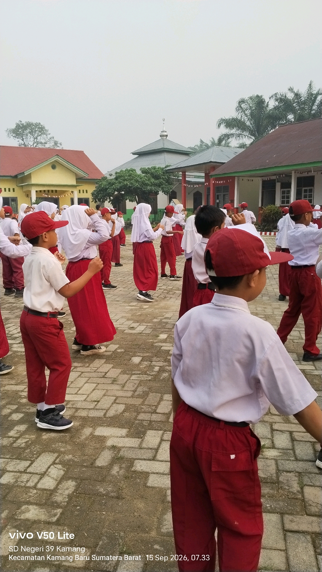 A group of elementary school children, most wearing red caps, white shirts, and red pants or skirts, are participating in an outdoor activity, possibly a physical education class or morning exercise, in front of school buildings. The setting appears to be in Indonesia, indicated by the presence of palm trees and architectural styles. Some children are facing away from the camera, engaged in coordinated movements, while others are looking towards the front. In the background, there are school buildings with red and yellow facades, a mosque with a green dome, and a cloudy sky. The ground is paved with interlocking bricks. Text at the bottom of the image indicates the device used is a "vivo V50 Lite", and it provides location information as "SD Negeri 39 Kamang, Kecamatan Kamang Baru, Sumatera Barat" and a date and time of "15 Sep 2026, 07.33".