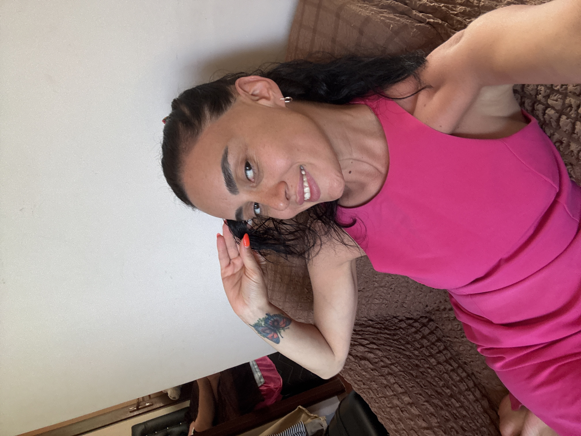 The image shows a woman with long, dark hair and fair skin, wearing a bright pink dress. She has a tattoo of a butterfly on her left forearm and is wearing hoop earrings. She is smiling and touching her hair. The woman is sitting on a brown, textured couch. In the background, a closet or wardrobe is visible with clothes hanging inside. The wall behind the couch is white. The lighting suggests an indoor setting, possibly a home. The scene does not provide any specific information that would allow for identification of the location within the city of Batumi, Georgia.