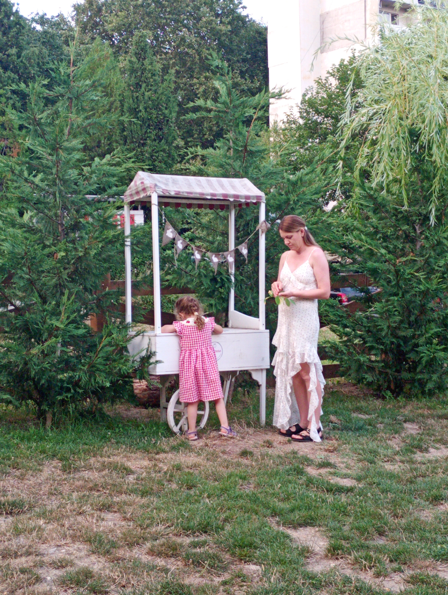 In Varna, Bulgaria, a woman in a white, ruffled dress stands next to a child in a pink checkered dress. They are beside a white cart with a red and white striped awning. The cart has a decorative banner with what appear to be animal figures hanging from it. The scene is outdoors, with lush green trees and dry grass surrounding them. A building is visible in the background, partially obscured by trees. The lighting suggests it is daytime, likely a warm day. The woman is holding a green leaf, and the child is leaning on the cart, looking down. The overall atmosphere seems calm and perhaps festive, given the cart's appearance.