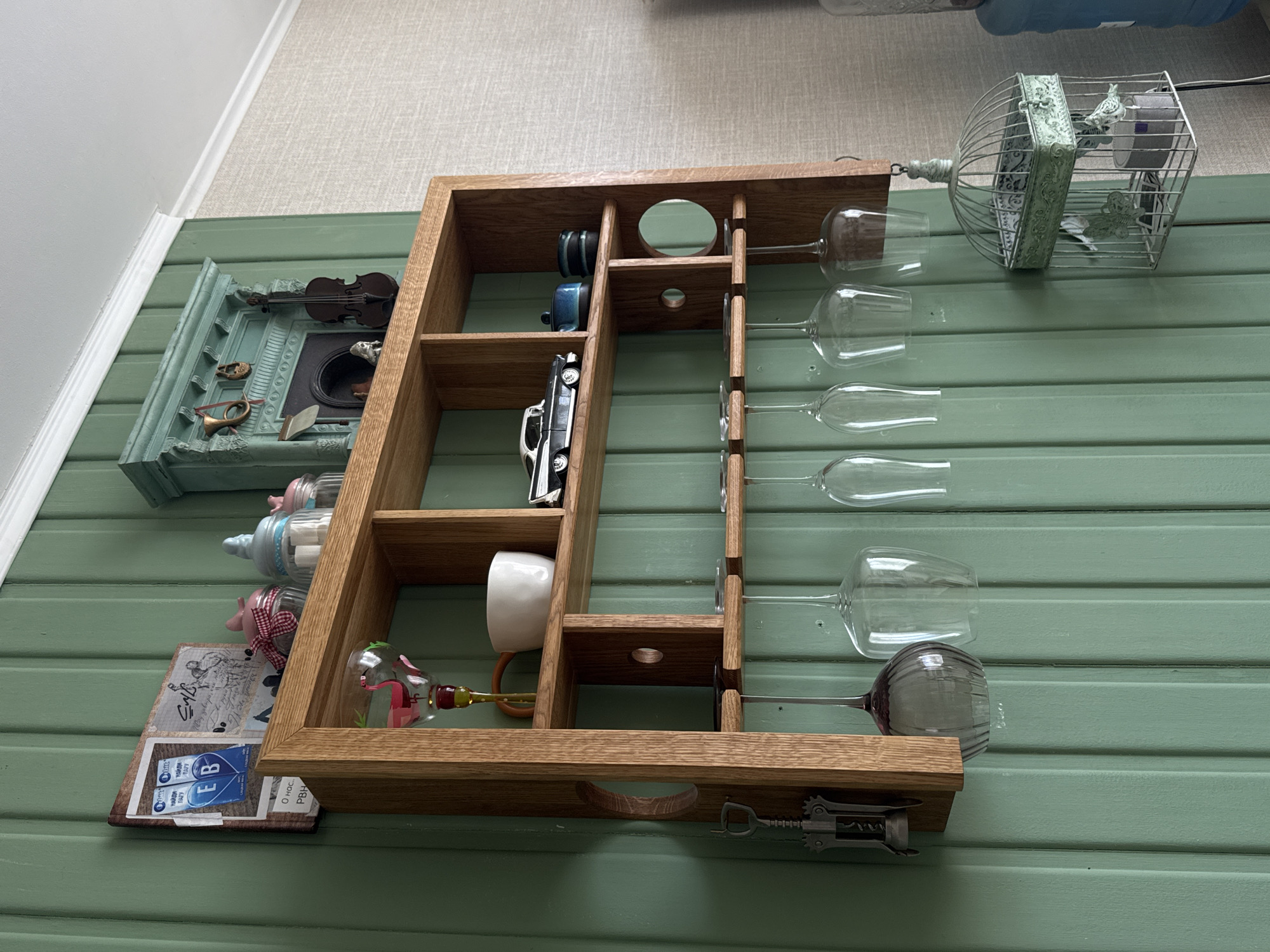 The image depicts a wooden shelf mounted on a pale green, vertically paneled wall. The shelf is L-shaped and features multiple compartments.
On the top shelf, from left to right, are two packets of something labeled "EB", a framed collage of images including what appears to be sheet music or drawings, a small glass jar with a red and white checkered lid filled with sugar cubes, and two other small jars with lids. To the right of these jars is a decorative turquoise miniature fireplace with various small objects placed on and within it, including a violin, a horn, and what appear to be two miniature shoes.
The middle shelf contains, from left to right, a decorative wine glass with a flamingo design, a white ceramic mug, a model of a vintage black and white car, and two small blue and black ceramic containers.
Below the middle shelf, a rack holds several wine glasses upside down. On the far left of the shelf's side, a corkscrew is attached.
To the right of the wine glasses, a decorative green birdcage is hanging. Inside the birdcage, there is a small bird figure and a cylindrical object.
The wall is painted a muted green and has distinct vertical paneling. The lighting suggests an indoor environment. There are no people visible in the image. The location is not specified as Kolomna, Russia.