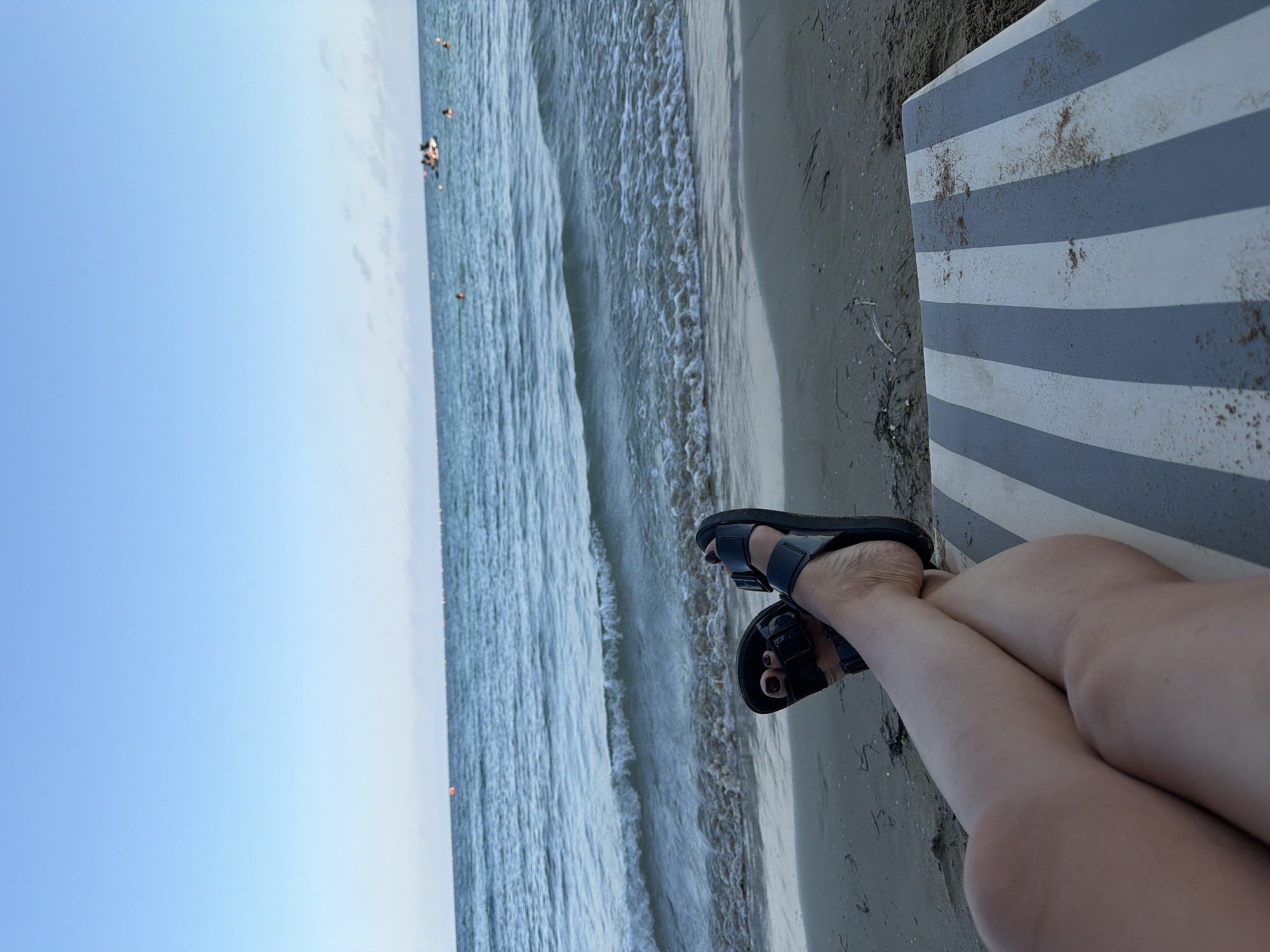 In Ravda, Bulgaria, a person's legs are seen crossed and resting on a striped beach chair. They are wearing black sandals. The person's feet are visible with painted toenails. The background shows the sea with gentle waves and a clear blue sky. A few buoys are visible in the water, and a small boat with people on it is further out at sea, along with some swimmers. The beach is sandy.