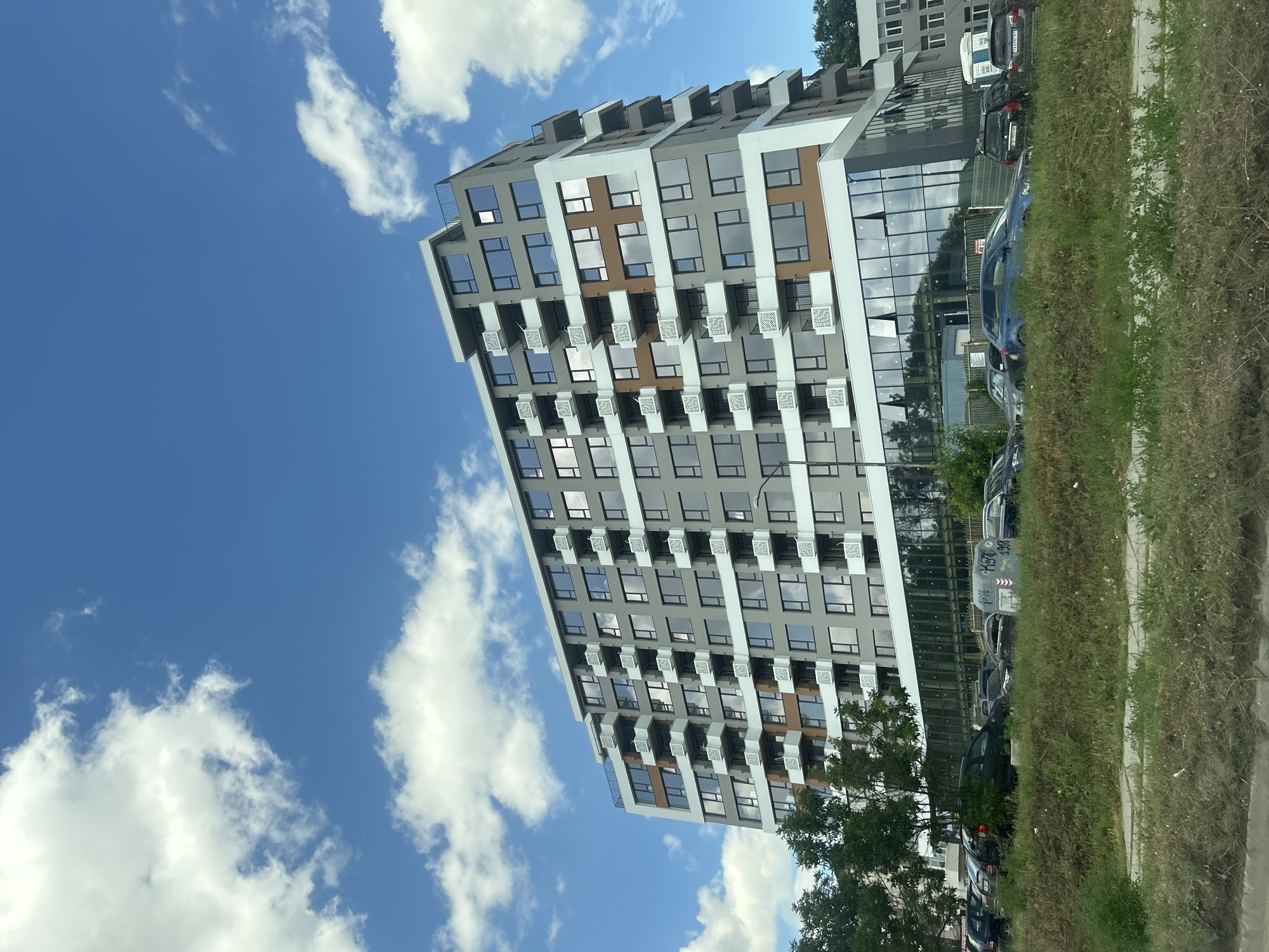 The image shows a modern, multi-story apartment building with a grey facade, white balconies, and accents of brown and white. The building has numerous windows and balconies, suggesting multiple residential units. In front of the building, there is a parking lot with several cars. The ground in front of the parking lot is covered with grass and some dry patches. In the background, trees and other buildings are visible. The sky is blue with scattered white clouds, indicating fair weather. The scene appears to be an urban or suburban environment.