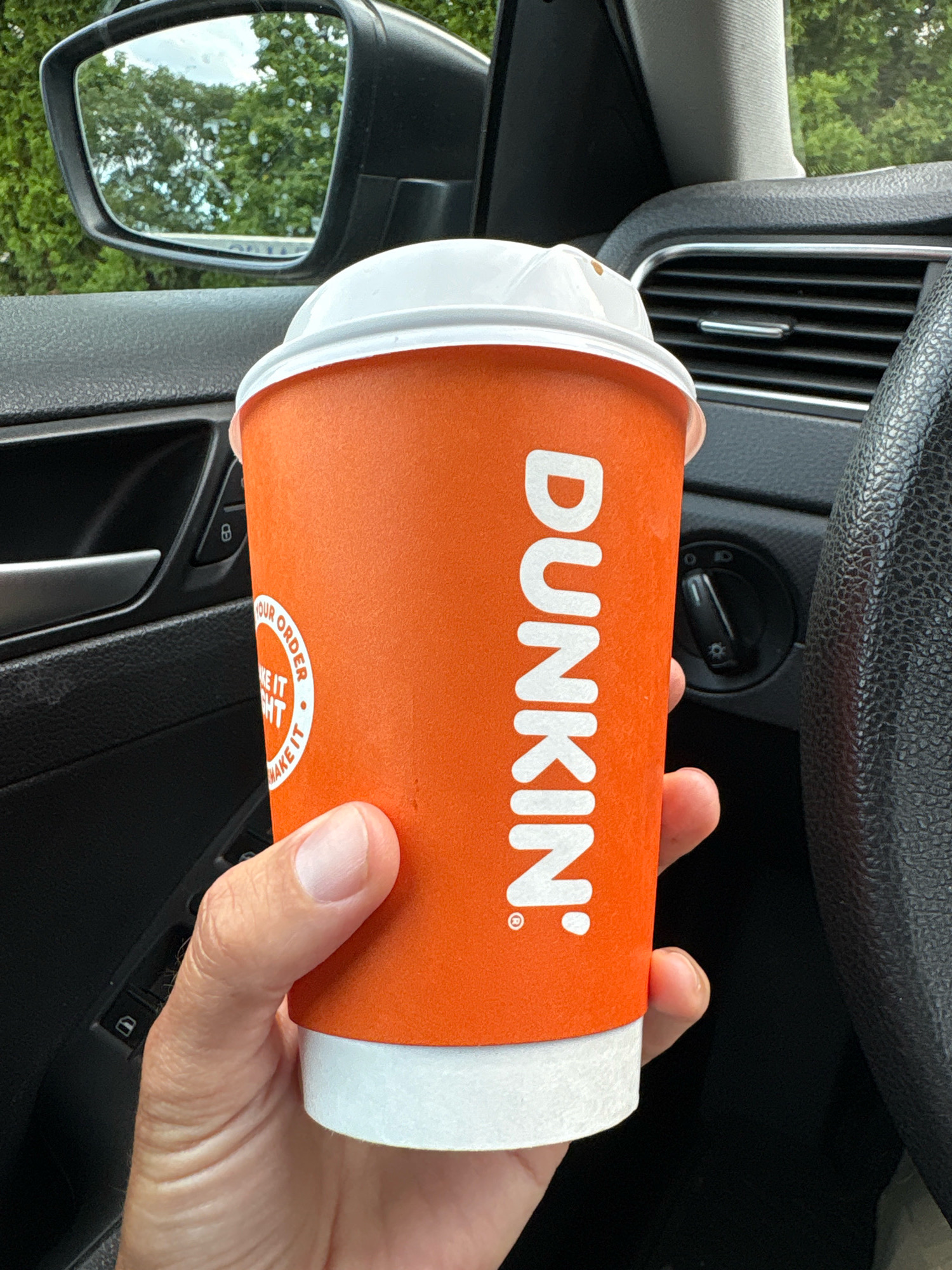 A person's hand is holding an orange Dunkin' coffee cup inside a car. The cup has a white lid and the Dunkin' logo is visible vertically on the side of the cup. The interior of the car, including the car door, a portion of the steering wheel, and an air vent, are visible. In the side mirror, trees and a cloudy sky are reflected. The image is taken from a first-person perspective.