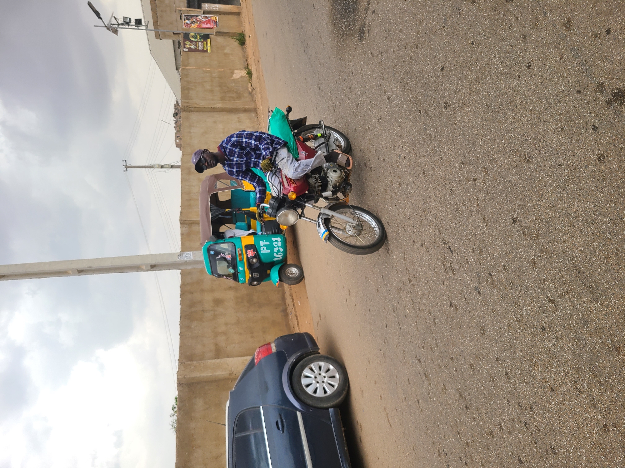 The image depicts a street scene in Jos, Nigeria. A man wearing a blue checkered shirt, cap, and glasses is straddling a red and chrome motorcycle. The motorcycle is parked near a blue "keke" or auto-rickshaw, which has the registration number PT-16321. A dark blue car is parked behind the keke. The street appears to be paved, and there is a concrete wall alongside the road. There is a utility pole in the background, and the sky is overcast. A building with advertisements on it can be seen in the distance.