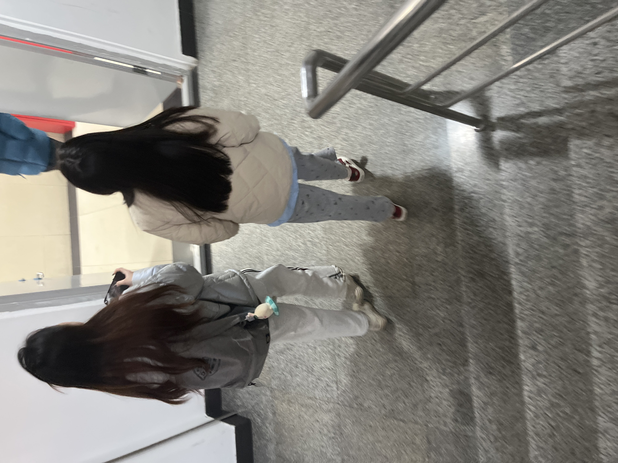 The image shows an overhead shot of two people standing on a gray speckled floor. The person on the right has long black hair, a beige puffer jacket, and patterned gray pants with red shoes. The person on the left also has long hair, and is wearing a gray jacket, gray pants with white stripes, and sneakers. A small toy is attached to the person on the left's jacket. A metal railing is visible in the upper right corner. Part of a white door or wall with a red accent is on the left side of the frame. The location is likely inside a building. The image was taken in Changchun City, China.