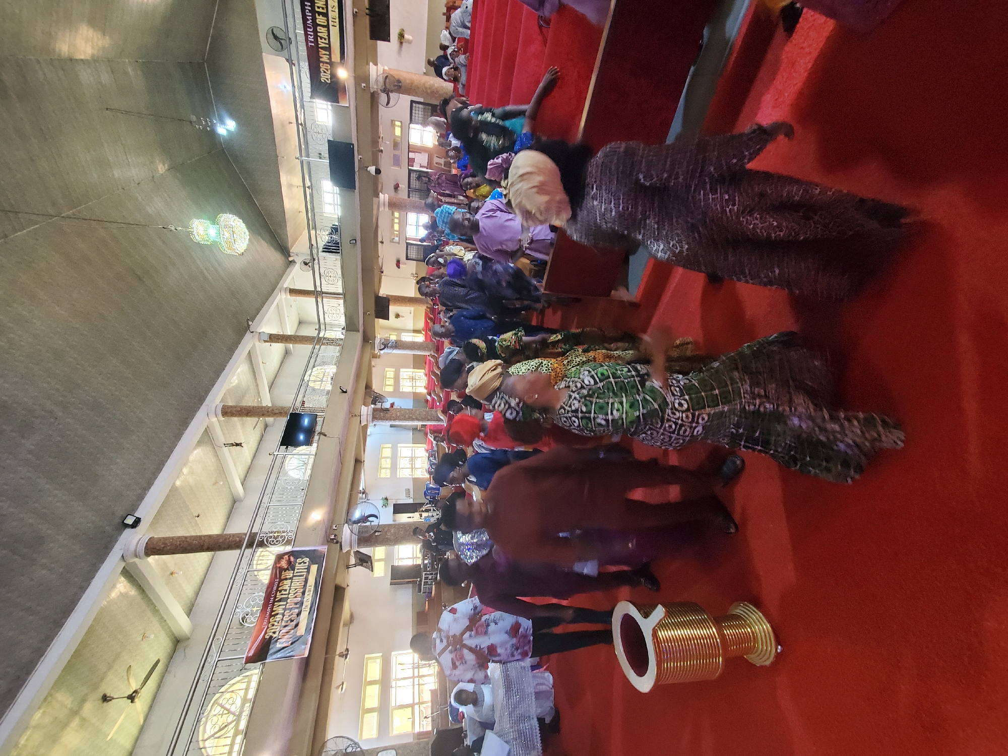 The image depicts a gathering in what appears to be a church setting, possibly in Onitsha, Nigeria. Numerous people, dressed in colorful attire, are present within a large hall with red carpeting. Some attendees are kneeling or prostrated on the floor, which suggests a moment of prayer or religious devotion. There is a large, golden structure in the foreground, along with a banner with the words "TRIUMPH IN CHRIST 2026 MY YEAR OF ENDLESS POSSIBILITIES". Chandeliers hang from the high ceiling.