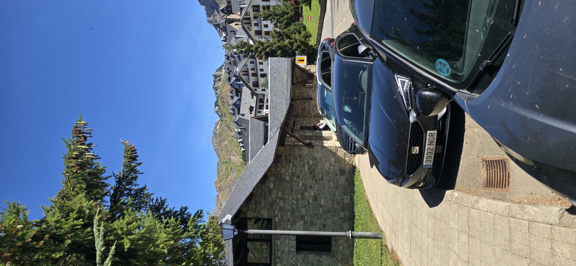 This is a vertical, eye-level, outdoor, daytime photograph taken in Sallent de Gállego, Spain. The foreground features the hoods of two black cars parked on a paved surface next to a sidewalk. The license plate of the closer car reads "3932 NGR". Behind the cars, a stone building with a slate roof is visible, with a lamppost on its left. Further back, a town with dense buildings and steeply pitched roofs stretches across a hillside, leading up to mountainous terrain under a clear blue sky. The architecture of the town suggests it is a ski resort. A few evergreen trees are scattered throughout the scene.