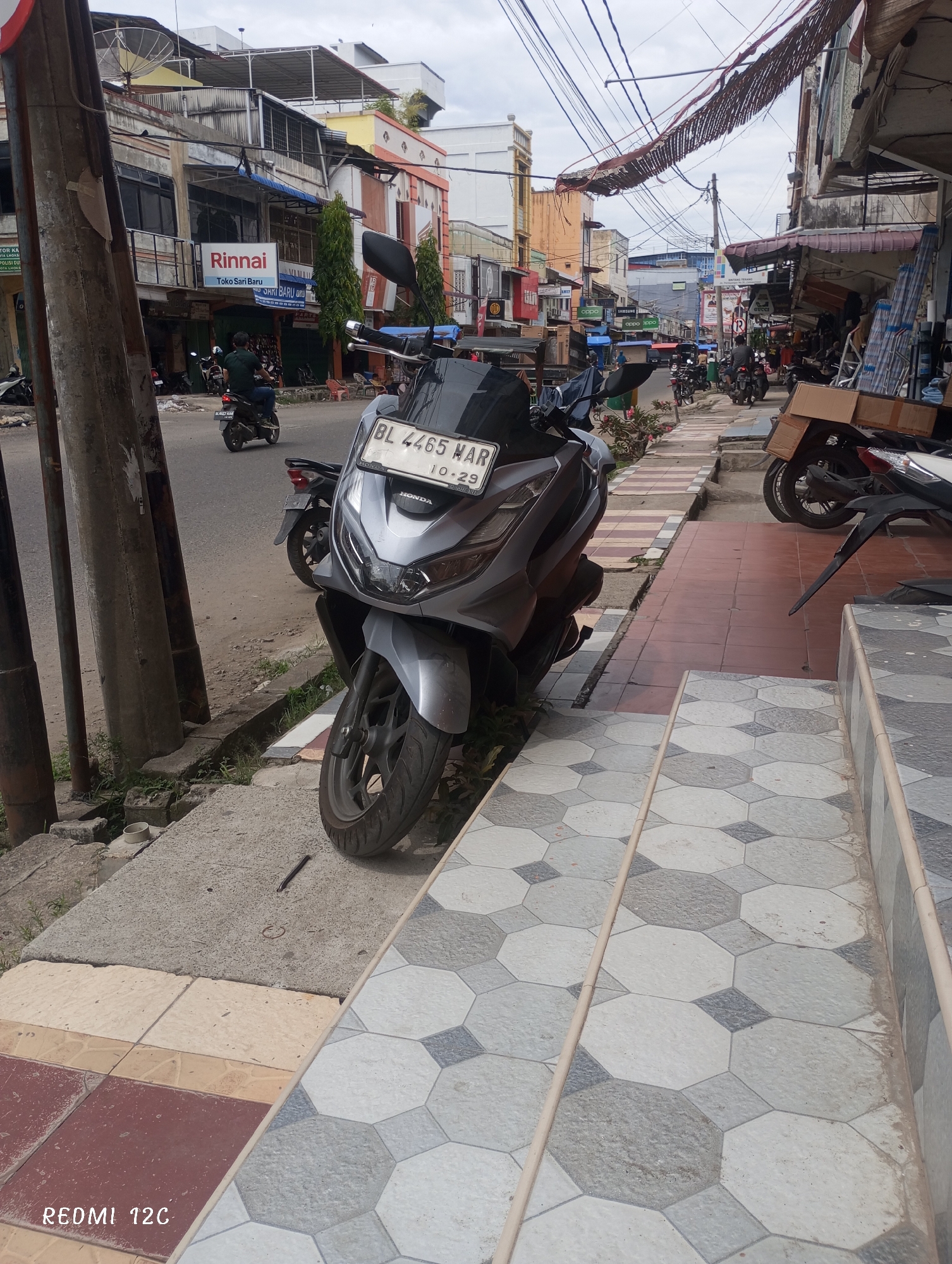 The image presents a street scene in Lhokseumawe, Indonesia. A silver Honda motorcycle is parked on the sidewalk, partially obstructing pedestrian access. The license plate reads "BL 4465 MAR" and indicates the date "10-29."
In the background, the street is lined with buildings, some showing signs of weathering or disrepair. Storefronts are visible, including a sign for "Rinnai Toko Sari Baru." Power lines and makeshift awnings crisscross above the street. Motorbikes are seen moving along the road. The sky is overcast. The sidewalk in the foreground features patterned tiles leading to steps.