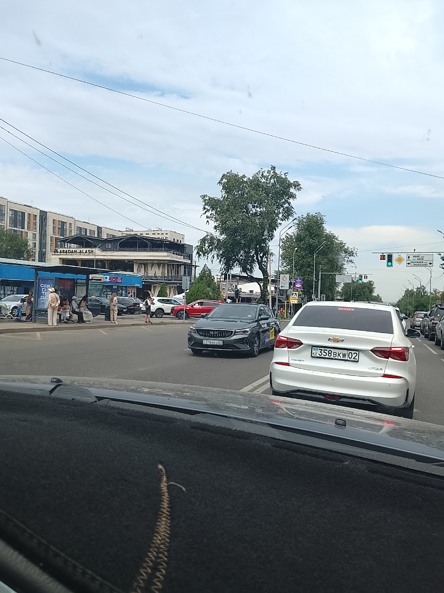 The image depicts a street scene in Almaty, Kazakhstan, likely during daylight hours with overcast skies. The foreground shows the interior of a vehicle, specifically the dashboard area covered with a dark, textured mat. In the midground, several vehicles are visible on a multi-lane road, including a white Chevrolet sedan with license plate "358BKV02" and a dark-colored sedan. A bus stop shelter with several people waiting is located on the right side of the road. Behind the bus stop, a multi-story building with "ABADAN-ALASH" signage is visible. Trees line the street, and power lines stretch across the sky. Traffic lights and street signs are also present. The overall atmosphere suggests a typical urban environment with moderate traffic and pedestrian activity.