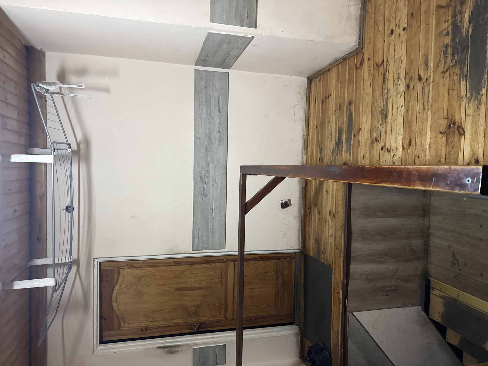 This is an indoor scene, likely a hallway or landing area, in Wąwelnica, Poland. The flooring is made of unfinished wooden planks with visible wear and some dark staining. A dark brown wooden railing runs diagonally across the foreground, obscuring a staircase descending into a lower level which has wood-paneled walls. To the left, a wooden door with a curved top is visible, set in a white frame. Black shoes are placed on a dark doormat to the left of the door. Above the door and extending across the scene is a metal clothes drying rack mounted to the wooden slatted ceiling. The walls are painted a light color and have horizontal decorative grey panels. There are some scuff marks and dark stains on the walls and floor.