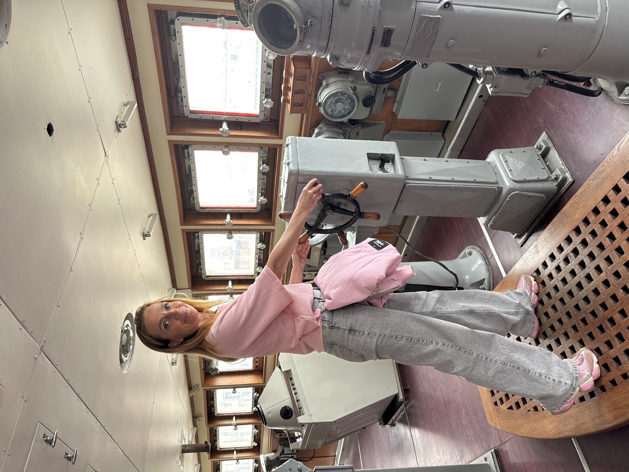 A woman stands in the bridge of a ship, her hands on the helm. She is wearing a pink t-shirt, gray jeans, and pink sneakers, with a pink backpack slung over her shoulder. The bridge is equipped with various navigational instruments, including a compass and a steering wheel. The room features large windows looking out onto a city, with wooden trim and white paneling. The floor is a dark reddish-brown, with a wooden grate in the foreground. This scene is likely from the central bridge of the Russian museum ship "Aurora" in Saint Petersburg, Russia, a historic vessel that played a significant role in the Russian Revolution.