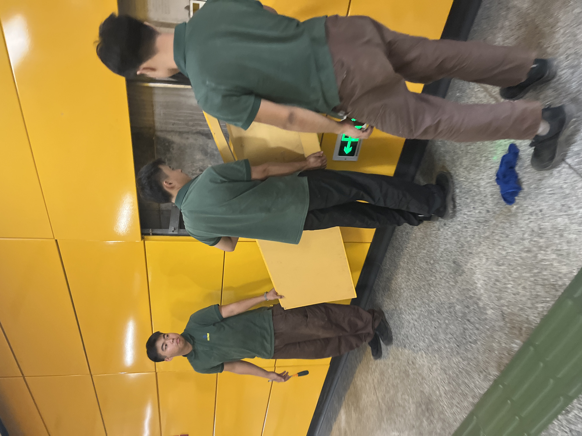 Three individuals in green polo shirts and brown or black pants are in front of a bright yellow wall in what appears to be a public transit station or similar enclosed area, possibly in Panyu District, China. The person on the left is holding a small orange screwdriver and a yellow rectangular object. The person in the middle is holding a larger yellow rectangular object, which is partially obscuring the wall behind them. The person on the right is facing away from the camera, also holding a yellow rectangular object. There is a green illuminated arrow sign pointing to the left on the yellow wall. The floor is a speckled gray material. A blue cloth is visible on the floor near the person on the right.