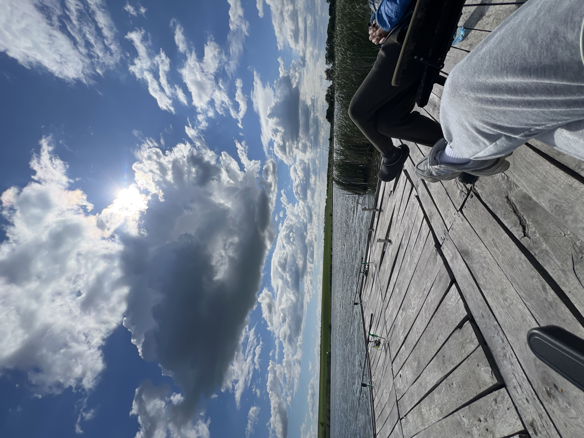 Two people are sitting on a wooden pier next to a body of water. Fishing rods are placed along the edge of the pier. One person is wearing dark-colored pants and shoes, while the other is wearing gray sweatpants and sneakers. The sky is blue with scattered white clouds, and the sun is visible. Grass and reeds line the opposite shore. The location is likely near a body of water in or around Kopychyntsi, Ukraine.