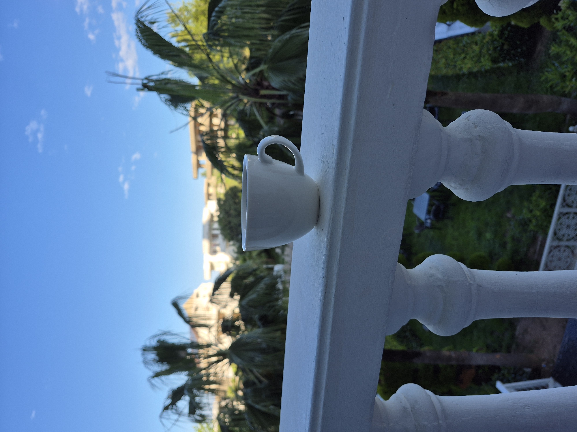 A white ceramic mug sits on a white painted balcony railing. Palm trees and a building can be seen in the background against a blue sky. The location is Alanya, Turkey.