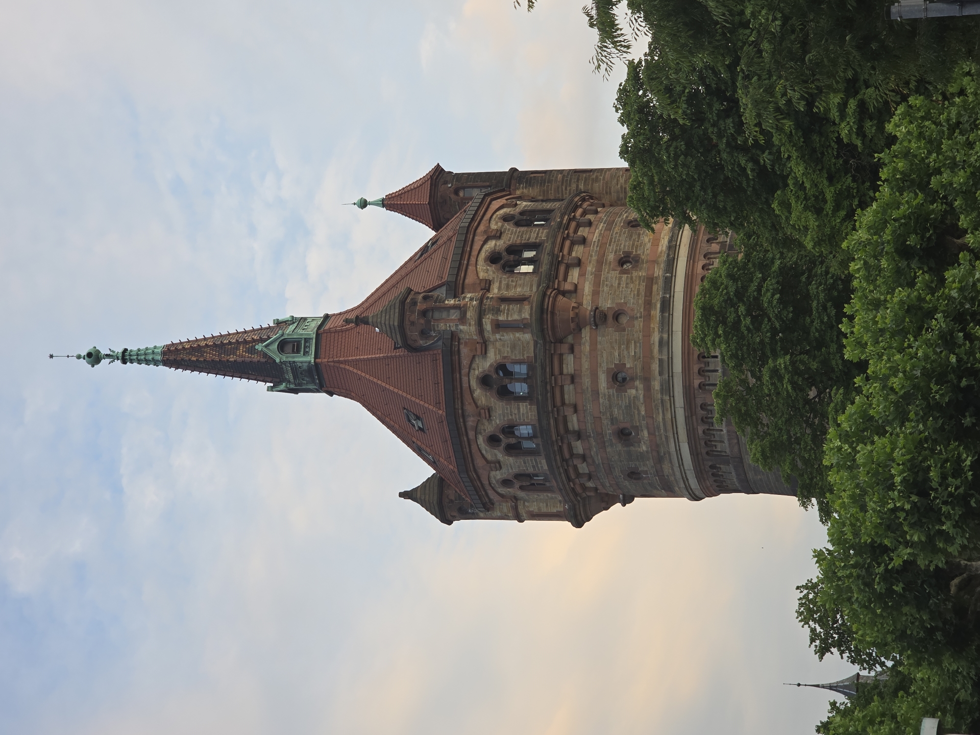 This is a vertical, eye-level, medium shot of the impressive Worms Water Tower, also known as the Wasserturm, in Worms, Germany. The tower dominates the frame, its reddish-brown and tan stonework contrasting with the overcast sky. The structure is octagonal with a conical, red-tiled roof topped by a green spire. Several smaller turrets with similar conical roofs flank the main tower. The tower has many arched windows and decorative stone detailing. Lush green trees and foliage partially obscure the base of the tower and extend into the lower portion of the image. A sliver of another spire, possibly part of a church, is visible peeking through the trees on the far left. The sky is a pale blue with scattered, soft clouds, tinged with the warm hues of either sunrise or sunset. No people are visible in the image.
