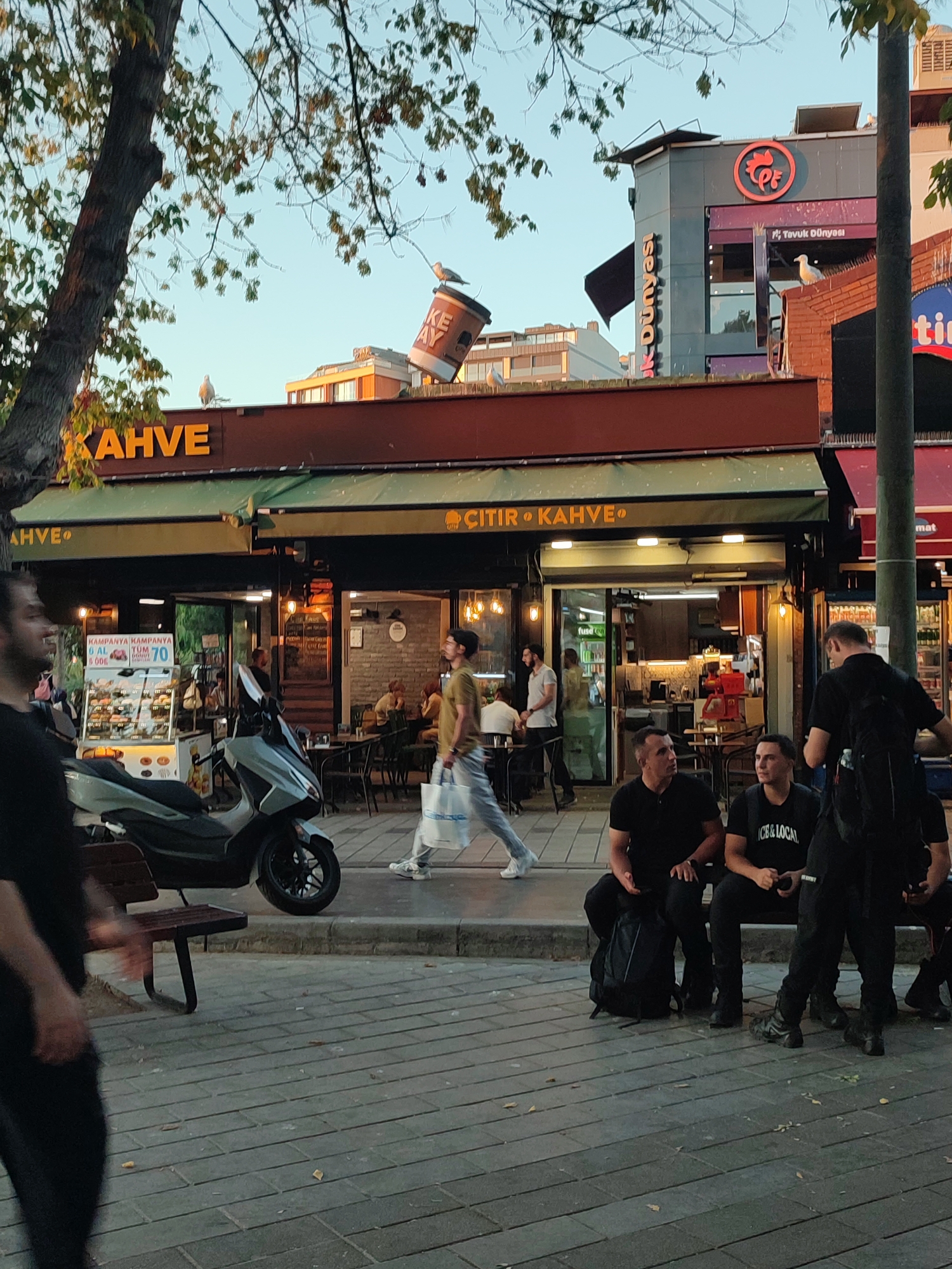 The image captures a street scene in Üsküdar, Turkey, with a focus on a cafe and its surroundings. A tree with autumn leaves is visible on the left, partially obscuring the sky. A large coffee cup prop is perched precariously on the roof of a building, with a bird on top of it and another bird on a ledge below. The cafe itself, branded with the word "KAHVE," features a green awning and outdoor seating. Inside, people are visible at tables.
In the foreground, a scooter is parked on the sidewalk, adjacent to a wooden bench. Several individuals are present on the sidewalk. Two men in black t-shirts are seated on the bench, facing the camera, while another man in black, carrying a backpack, stands to their right. Further down the sidewalk, a person in a black shirt and black pants is walking away from the camera, their face blurred. Another individual is walking towards the right of the frame, carrying a white shopping bag. A display stand for food items is positioned to the left of the cafe entrance, with a sign advertising a special offer. The street is paved with grey tiles, and fallen leaves are scattered across the surface. In the background, other buildings and signs are visible, including a sign for "TAVUK DÜNYASI" (Chicken World). The lighting suggests it is late afternoon or early evening, with a warm, soft glow.