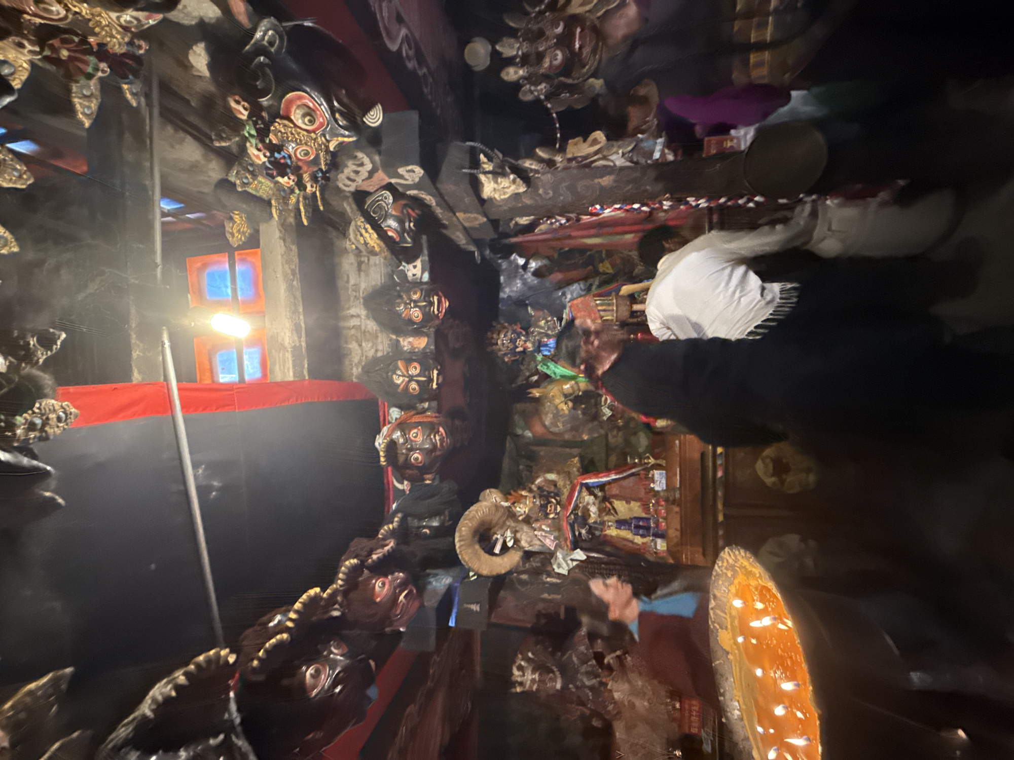 The image captures a dimly lit, richly decorated interior, likely a temple, shrine, or ceremonial space. Numerous stylized masks or sculptural heads adorn the walls and ceiling beams, many displaying fierce or elaborate expressions, some with horns or intricate gold and red details. Several masks are dark-colored with prominent red eyes, contributing to a mystical or imposing atmosphere.
Several people are present, mostly seen from the back or side, suggesting they are observing or participating in an event. One individual, wearing a white shirt and dark trousers, stands prominently in the middle ground, facing deeper into the space. Another person, partially visible in the right foreground, appears to be wearing dark clothing.
Among the various objects and decorations, a ram's skull with large, curved horns is visible on a wooden beam. In the foreground, a long, boat-shaped vessel holds numerous glowing lights, possibly candles or oil lamps, casting a warm, orange illumination. Colorful fabrics, including red and striped patterns, are draped or hung throughout the space, along with what appear to be offerings or decorative items on shelves. A bright, rectangular artificial light fixture is visible on the left.
The scene conveys a sense of solemnity or reverence, indicative of a religious or cultural gathering. The distinct style of the masks and decorations strongly suggests a cultural context from the Himalayan region, such as Tibetan Buddhism. The indoor setting is lit by artificial light sources, implying it could be evening or a space with limited natural illumination. No discernible text is visible.