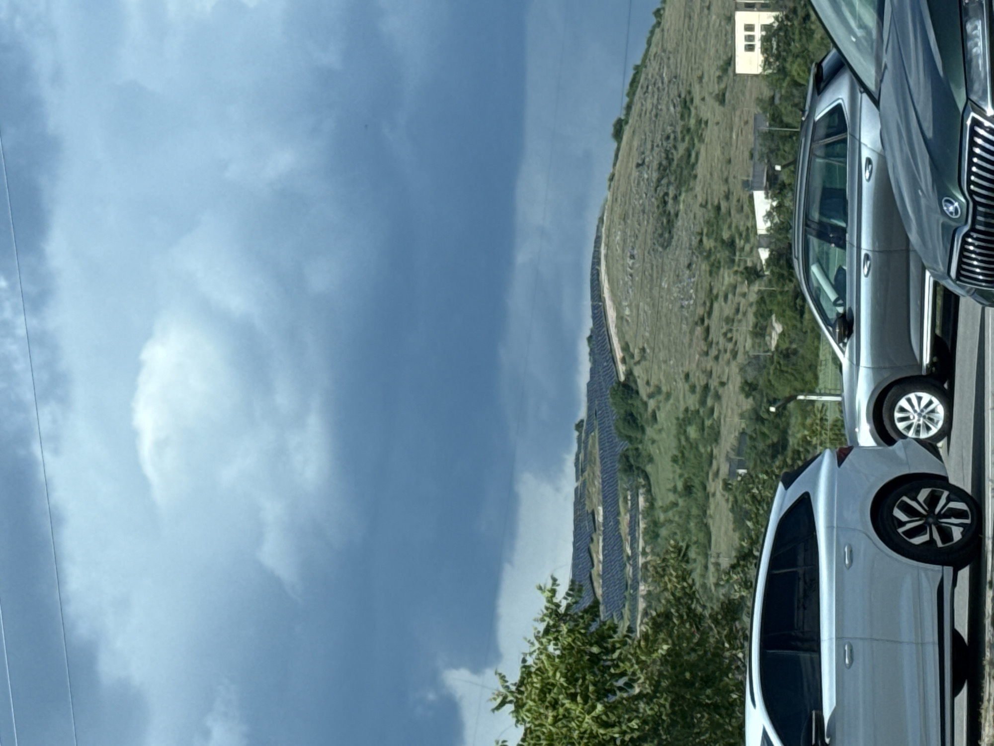 The image shows a view from the city of Stara Zagora, Bulgaria, featuring a partly cloudy sky with a section of the landscape that includes a hillside covered in green vegetation and a solar panel array. A couple of cars are parked nearby; one is light-colored, and the other is gray/silver. A building is seen in the distance, potentially a residence.