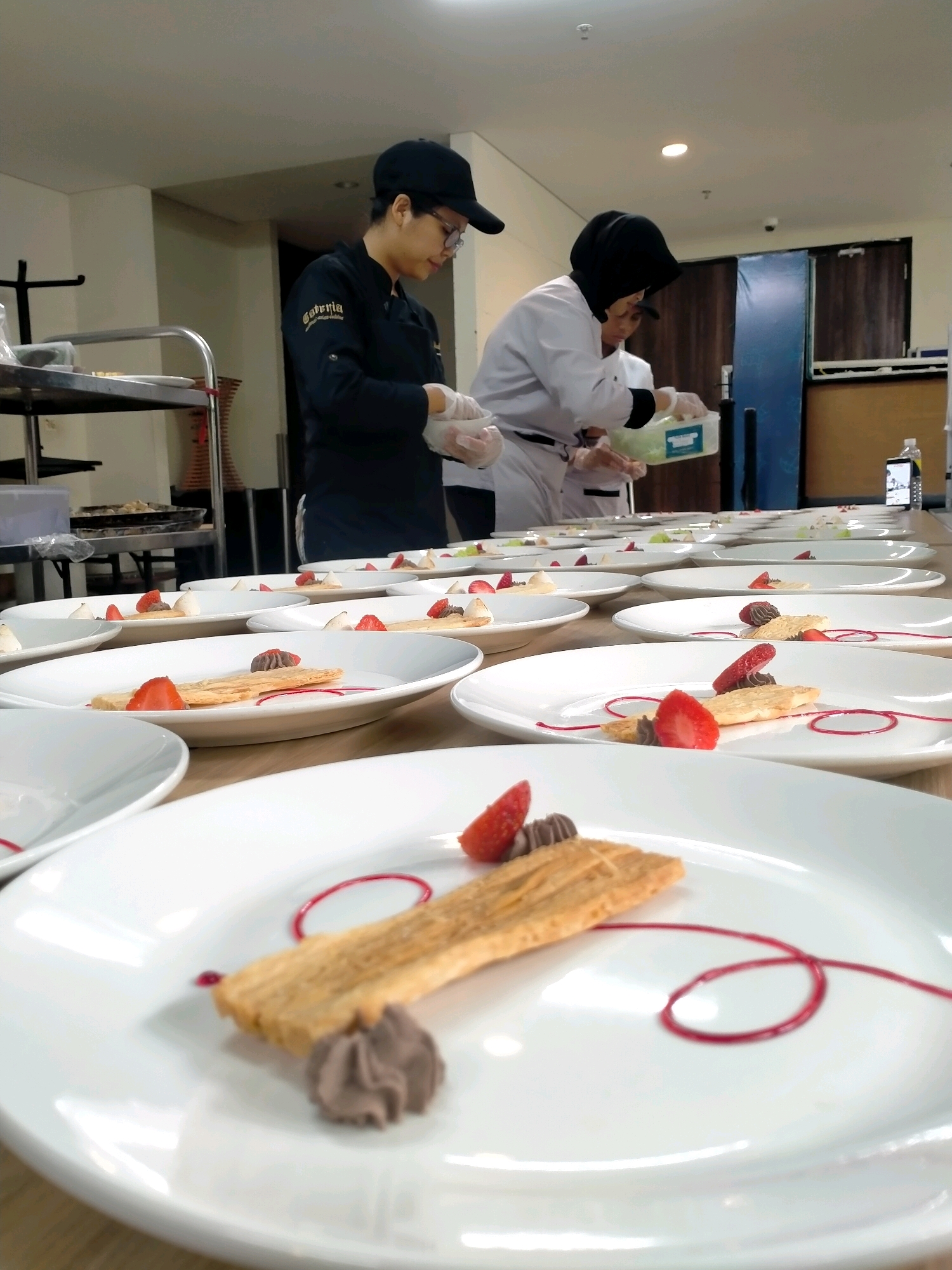The image shows a group of people in a kitchen or food preparation area, likely in Batu, Indonesia. Two individuals are visible in chef's uniforms. One person on the left wears a black chef's jacket and a black baseball cap, with glasses. They are wearing gloves and appear to be handling a white bowl. The second person, to the right, is wearing a white chef's jacket and a black hijab. This person is also wearing gloves and is holding a clear plastic container filled with green ingredients, possibly salad or herbs.
The foreground and midground of the image are filled with multiple white plates, each meticulously arranged with a dessert. The desserts appear to consist of a thin, elongated cracker or biscuit, a dollop of chocolate mousse, slices of fresh strawberries, and a swirl of red sauce. Some plates also feature small meringues.
In the background, a stainless steel cart is visible on the left, laden with more white plates. Further back, there are cabinets and doors, suggesting a professional kitchen environment. The lighting is bright, with ceiling lights visible. The overall scene depicts a team of chefs preparing plated desserts for service.