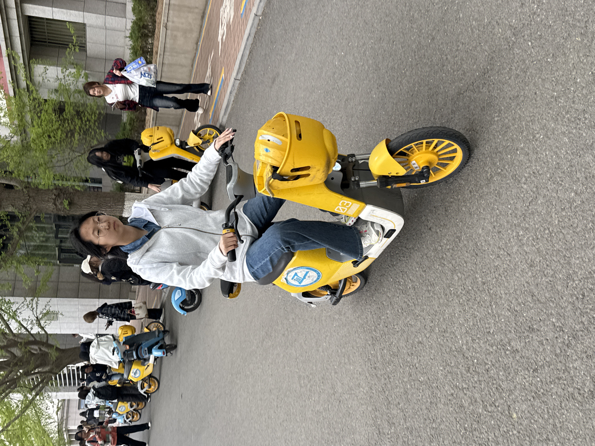 The image captures an outdoor scene, likely a paved pathway or street in a campus or urban area, bustling with people and shared electric scooters during the daytime.
In the foreground, a young woman with glasses and dark hair is seated on a bright yellow electric scooter, gripping the handlebars. She is wearing a light grey hooded jacket over a blue denim top, blue jeans, and white sneakers with green accents. She looks towards the viewer with a neutral expression. The scooter she is on features a yellow body, a black seat, and a distinctive yellow front basket with a slightly anthropomorphic design, showing a "B" in a blue circle and "03" on its side. Another blue circular logo is visible on the scooter's body near the seat.
Behind her, several other individuals are present. To her right, a woman with long dark hair, wearing a black top, is also on a yellow scooter. Further back, another woman in a plaid shirt and dark jeans is walking, holding a white bag with blue text and appearing to look at her phone. In the mid-ground and background, a line of similar yellow and some blue electric scooters are parked or being ridden by other people, some walking alongside. The scene suggests a popular shared mobility service.
The pathway is asphalt, and to the left, a brick-patterned path runs alongside. Buildings with windows are visible in the background, and trees with fresh green foliage indicate a spring or summer setting. The lighting is diffused, suggesting an overcast day or indirect sunlight. The overall atmosphere is casual and active, typical of a busy public space.