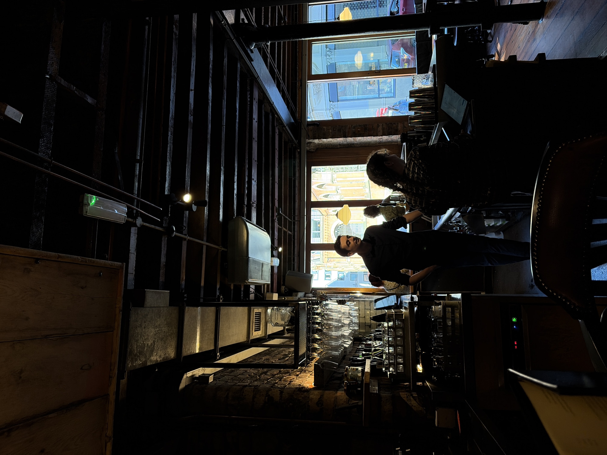 The interior scene shows the dimly lit inside of a bar. Two people are partially visible, with one man standing behind the counter. Behind the counter, multiple clean glasses hang upside down. The room's decor has wooden details and pipes along the walls. Sunlight streams through the windows, where brick buildings, flags, and street details can be seen.