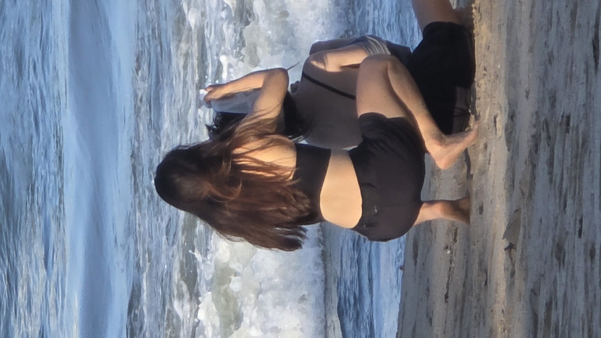 Two individuals are standing on a sandy beach at Nha Trang Ward, Vietnam, where the ocean water is flowing. One individual is closer to the camera, has long dark hair, and is wearing a black crop top and black shorts. The second individual is wearing a gray shirt and dark shorts. Both individuals appear to be interacting with the ocean water.