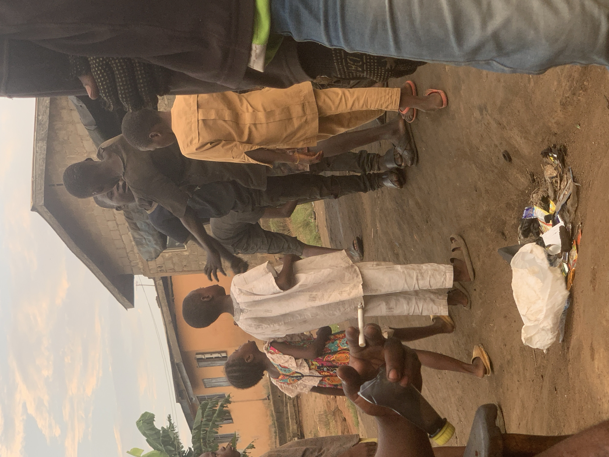 In Shagamu, Nigeria, a group of people, including several children, are gathered outdoors on a dirt ground. The sky above is a pale orange, suggesting either sunrise or sunset. In the foreground, a person's hand holds a lit cigarette. Several individuals are dressed in traditional clothing. One child, in a white outfit, stands facing away from the camera, while another child in a colorful dress is seen from the side. Behind them, a few adult males are interacting. In the background, there are low-rise buildings with corrugated iron roofs. The ground is littered with various pieces of trash, including plastic bags and wrappers. The overall scene depicts a moment of everyday life in a Nigerian community.