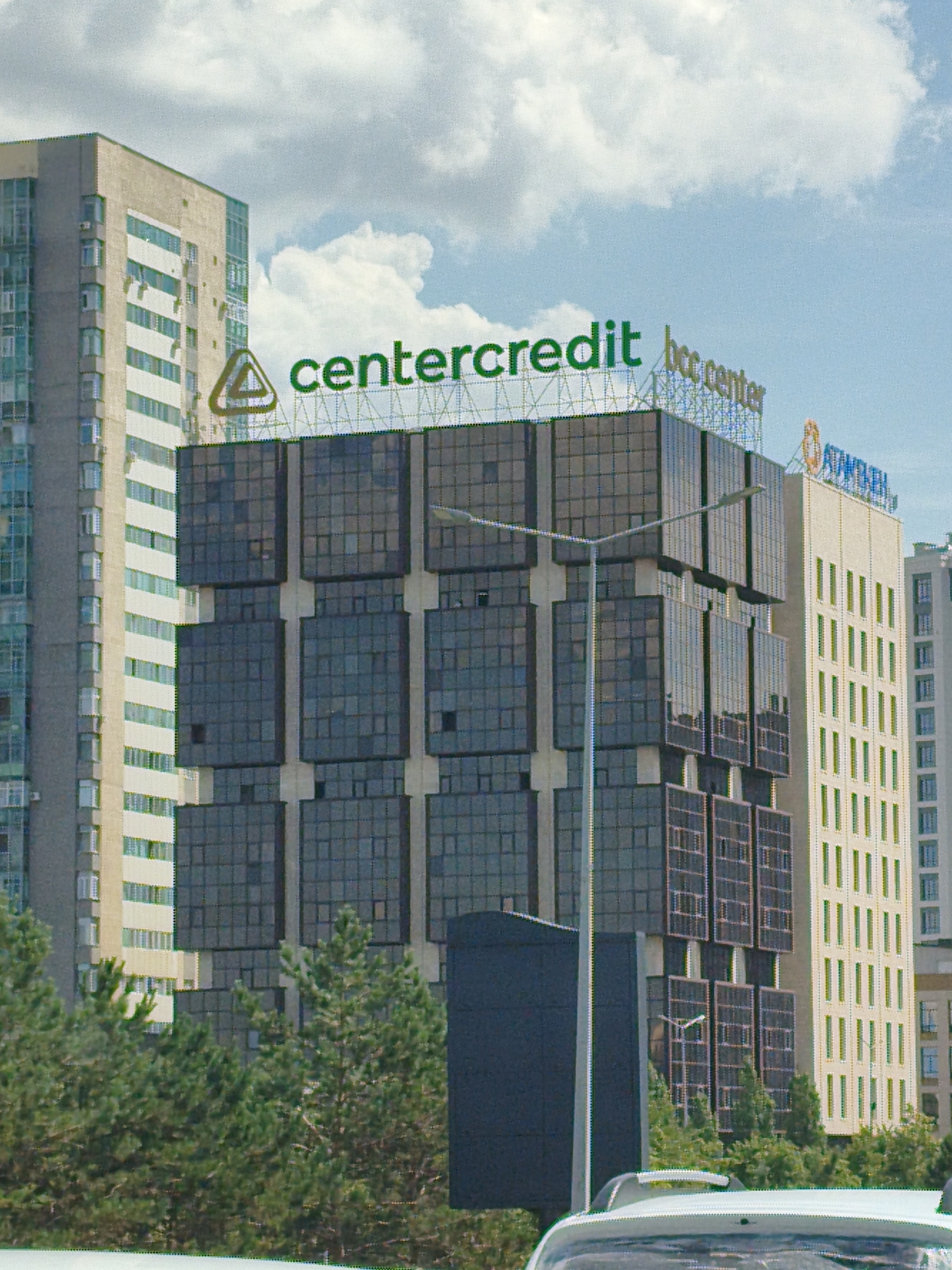 The image depicts a street scene in Astana, Kazakhstan, featuring several modern buildings. The primary focus is on a large, dark-paneled building with a prominent green sign reading "centercredit" and a smaller sign "bcc center." To the left, a lighter-colored, multi-story building with numerous windows is visible. To the right, another light-colored building with a distinctive yellow facade and rows of windows stands tall. The buildings are set against a backdrop of a partly cloudy sky. Trees line the lower portion of the image in front of the buildings. A lamppost is positioned in front of the "centercredit" building, and the roof of a white vehicle is visible at the bottom of the frame. The overall impression is one of urban architecture and city life.