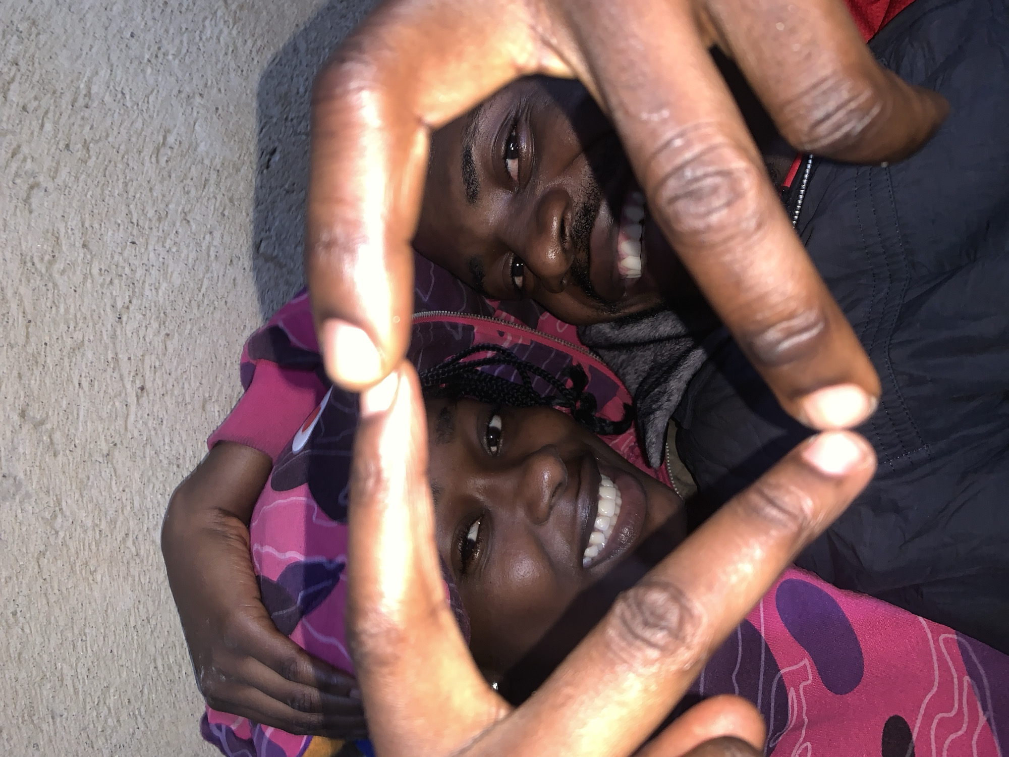 The image is a close-up, overhead, indoor shot of two young Black adults, a male and a female, lying down. They are forming a heart shape with their hands in the foreground, with their faces visible behind their joined hands. The woman is on the left, smiling and looking at the camera. She has braids and is wearing a pink and purple patterned garment. The man is on the right, also smiling and looking at the camera. He has a mustache and goatee and is wearing a dark jacket with red trim. The background appears to be a textured wall, possibly concrete, with a shadow cast on it from the subjects. The overall lighting is dim, suggesting an indoor setting, possibly at night or in a dimly lit area. The context of Rafin Par, Nigeria is not evident from the visual information in the image itself.