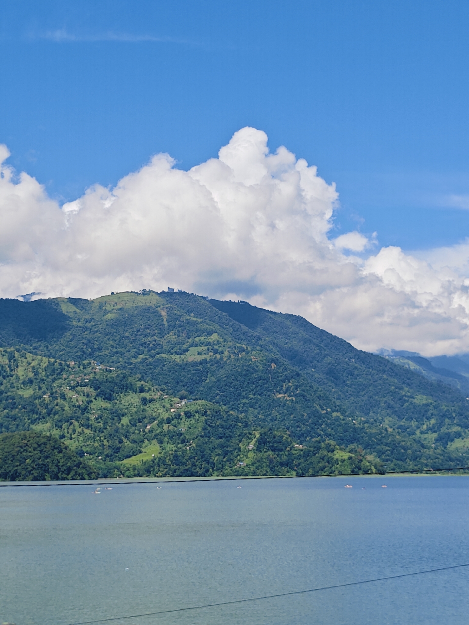 This is a landscape photograph of Phewa Lake in Pokhara, Nepal. The foreground is dominated by the tranquil, blue water of the lake, with a few small boats scattered across its surface. In the middle ground, a lush green, tree-covered mountain rises dramatically from the water's edge. The mountain slopes are dotted with small settlements. The background features a vibrant blue sky with large, fluffy white clouds. The overall impression is one of natural beauty and serenity.