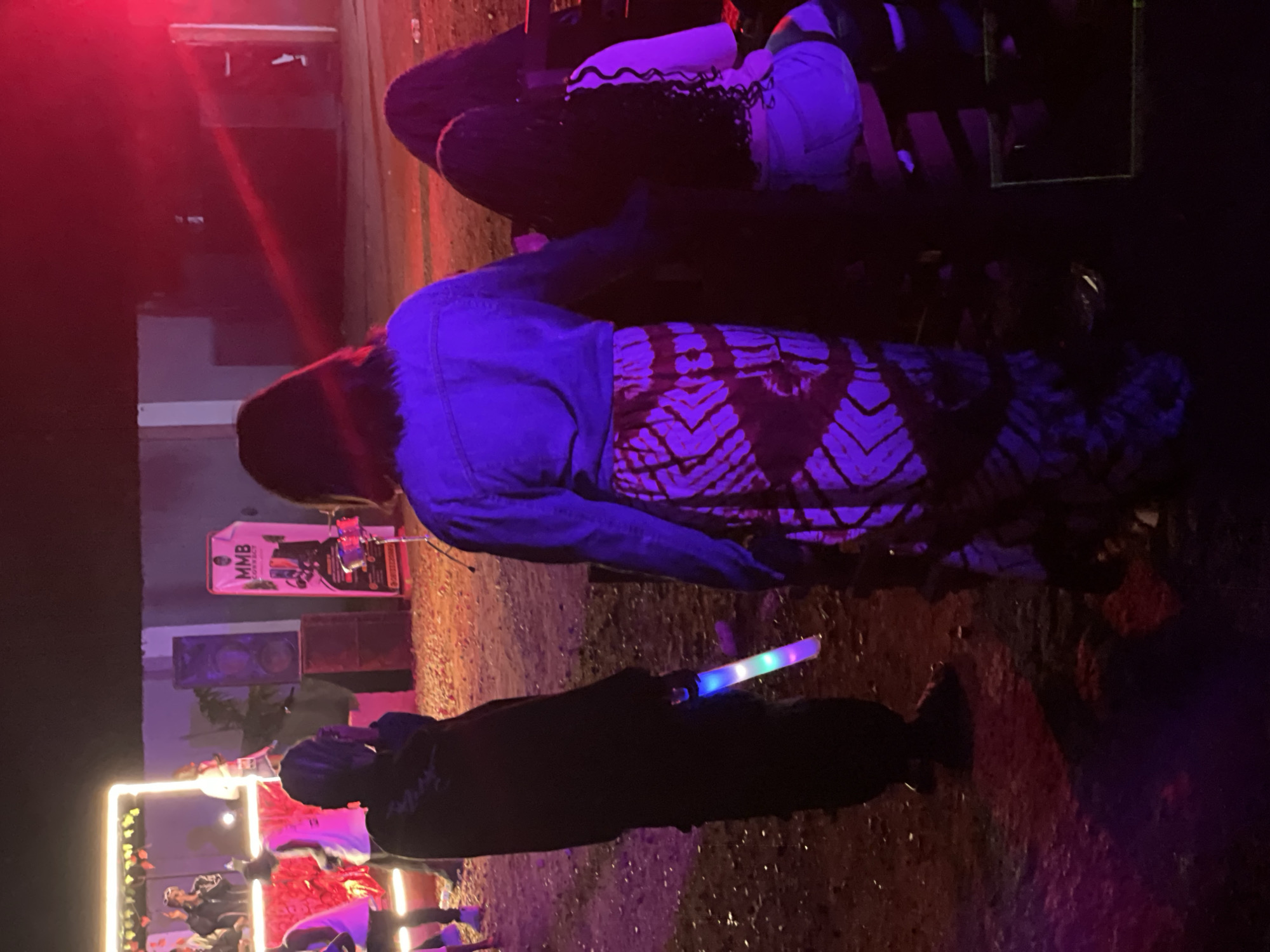 In the city of Durumi, Nigeria, at what appears to be an outdoor event, a group of individuals are gathered. The scene is illuminated by a combination of artificial lighting, including neon lights outlining a raised platform and bright red stage lights. The ground is covered in gravel and dirt.
In the foreground, a person wearing a black outfit is holding a multi-colored glow stick. To their right, another individual is visible from the back, wearing a denim jacket and a patterned skirt. Further in the background, several other people are present, some standing and some seated on benches. A banner with "MMB CONNECT" printed on it is propped up near some speakers. The overall atmosphere suggests a lively gathering, possibly a concert or a party.