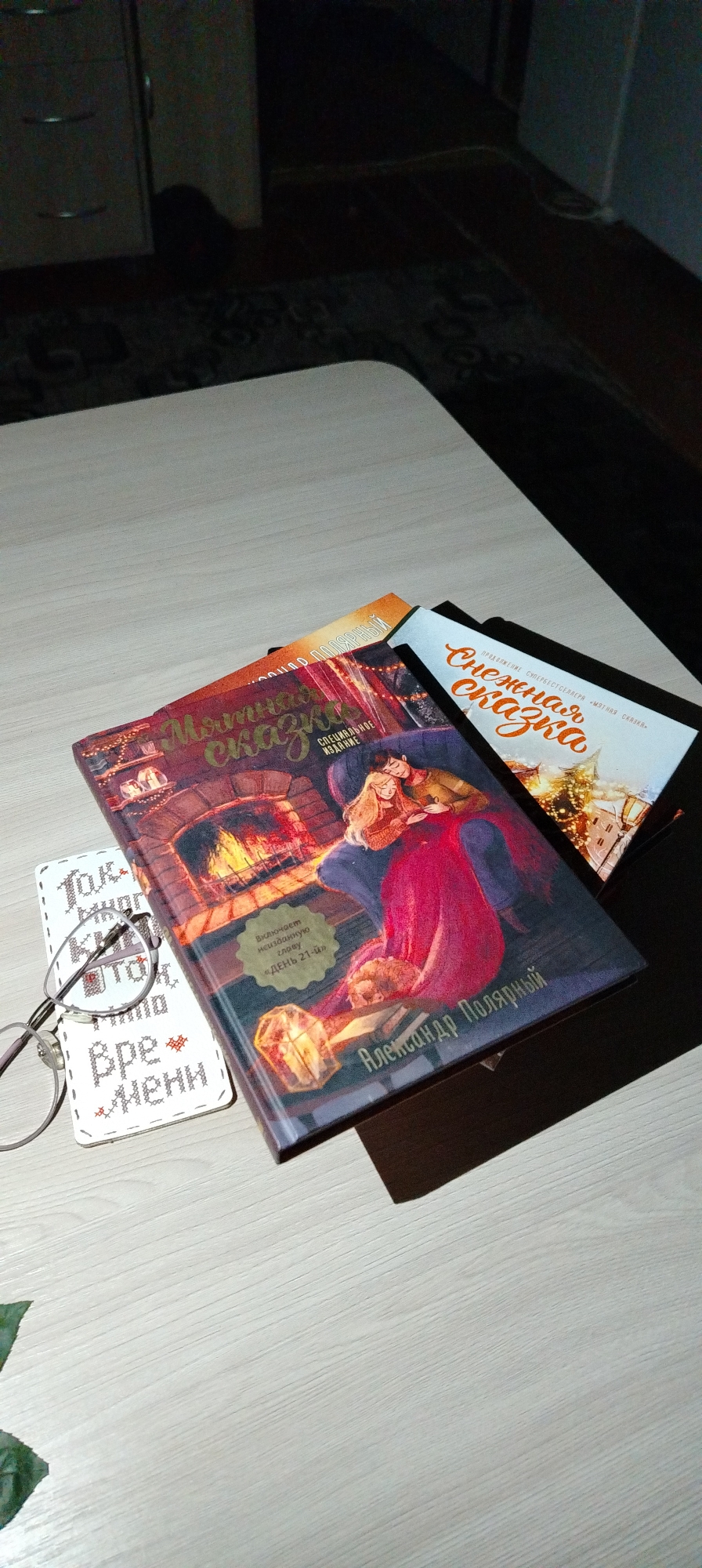 A top-down, slightly high-angle shot shows a light-colored, possibly wooden table or surface in an interior setting. Three books are arranged on the table: "Мятная сказка" (Mint Tale) by Александр Полярный (Alexander Polar), "Снежная сказка" (Snow Tale), and an orange book. "Мятная сказка" is on top, its cover featuring an image of a couple sitting by a fireplace. Next to the books lies a pair of eyeglasses and a small rectangular cross-stitched object. Part of a dark cabinet or furniture is visible in the background. There are no people in the frame. The location, based on the books' author and language, is likely within Russia. The city of Chaikovsky is not visible, and there is no way to determine its relevance to this picture.