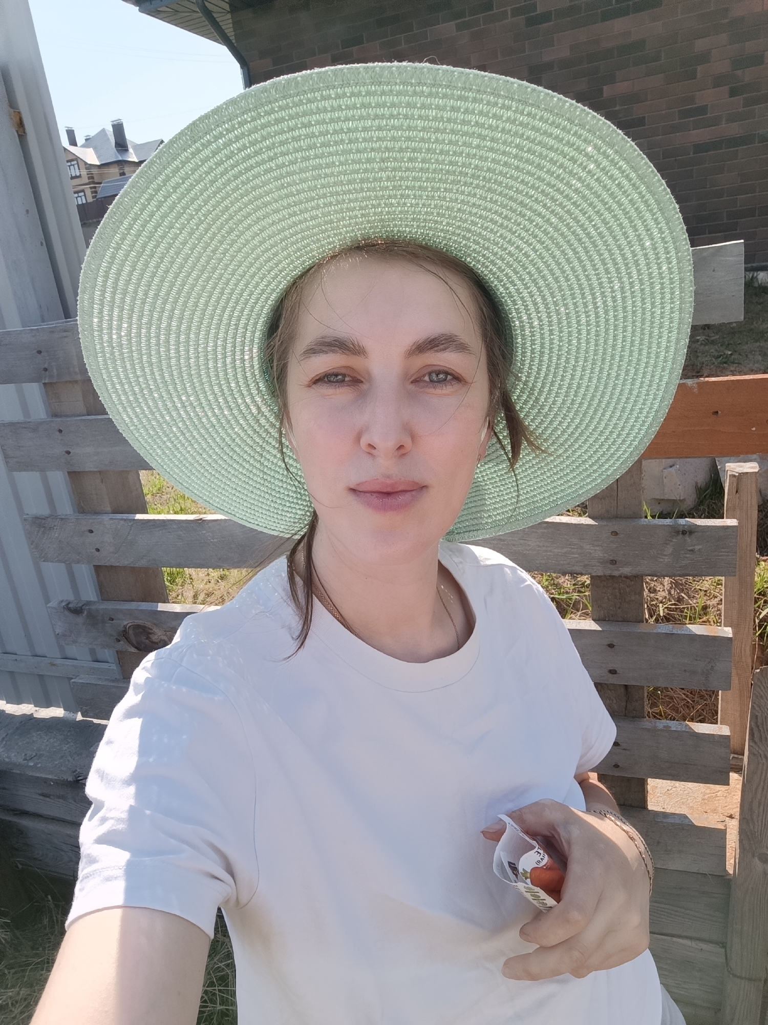 A woman with fair skin and light brown hair is the subject of a medium shot, set outdoors in Chaikovsky, Russia. The woman's hair is pulled back, and she is wearing a large, pale green sun hat and a plain white t-shirt. She holds a small package with one hand. A weathered wooden fence acts as the backdrop, with hints of green grass and foliage visible between the slats. In the distance, brick buildings and a grey roof can be seen. The lighting suggests a bright, sunny day.
