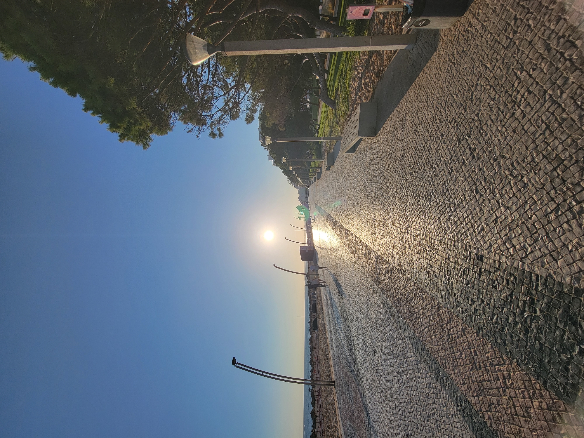 The image depicts a sunlit promenade in Quarteira, Portugal, likely at sunrise or sunset given the low angle of the sun. The promenade is paved with cobblestones that reflect the sunlight, creating a shimmering effect. To the left, a beach with numerous sun umbrellas stretches towards the horizon, meeting the calm sea under a clear blue sky. Several modern, curved lampposts are spaced along the promenade.
In the middle ground, a few individuals are walking, casting long shadows on the cobblestone path. The path continues into the distance, lined with more lampposts and benches. To the right, a line of trees with dense green foliage borders a grassy area. Another lamppost, taller and more utilitarian, stands closer to the foreground on this side, next to a waste bin and a small sign. The overall scene conveys a sense of tranquility and early morning or late evening activity in a coastal city.